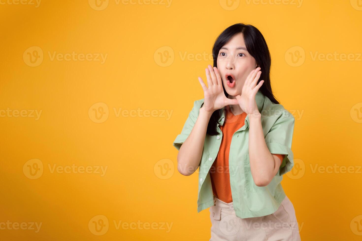 Cheerful Asian woman 30s wearing orange shirt and green shirt showing hand gesture covering mouth, symbolizing shout or surprise. Energetic moments and conveying sense of exuberance concept. photo