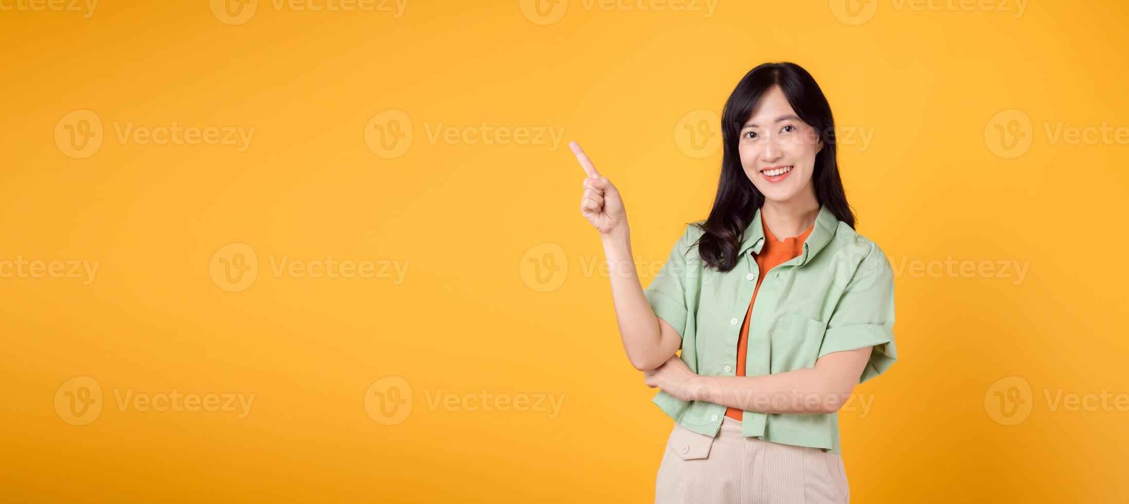 young 30s Asian woman wearing a green shirt on an orange shirt, pointing with excitement to free copy space. Discover the concept of discount shopping promotion with captivating image. photo