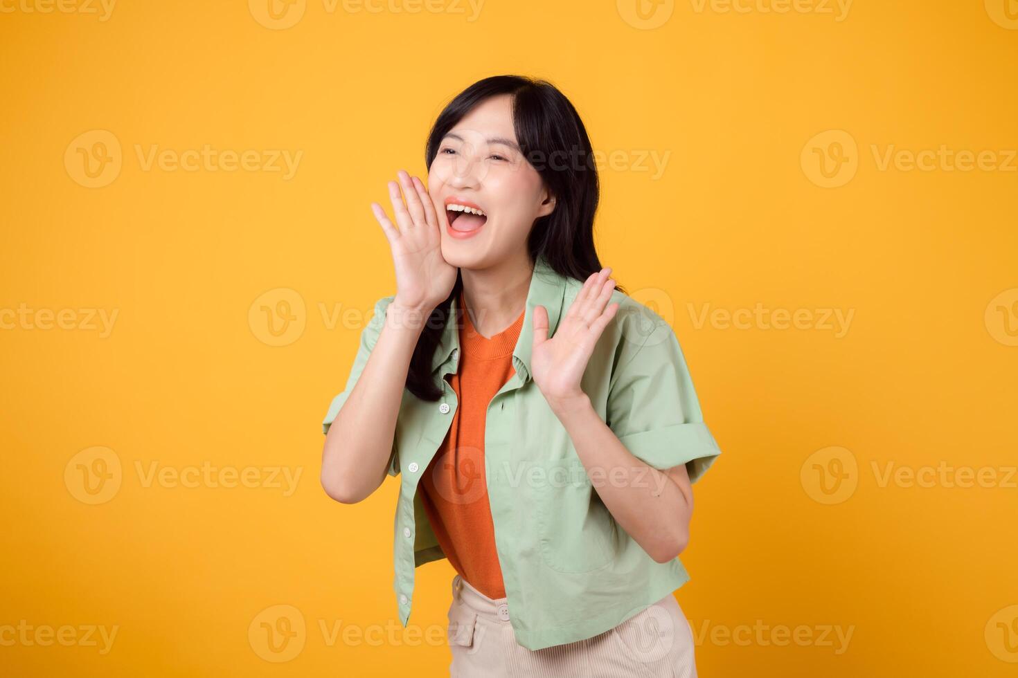Energetic young Asian woman in her 30s wearing a green shirt on an orange background, shouting with excitement. Explore the concept of discount shopping promotion with this vibrant image. photo