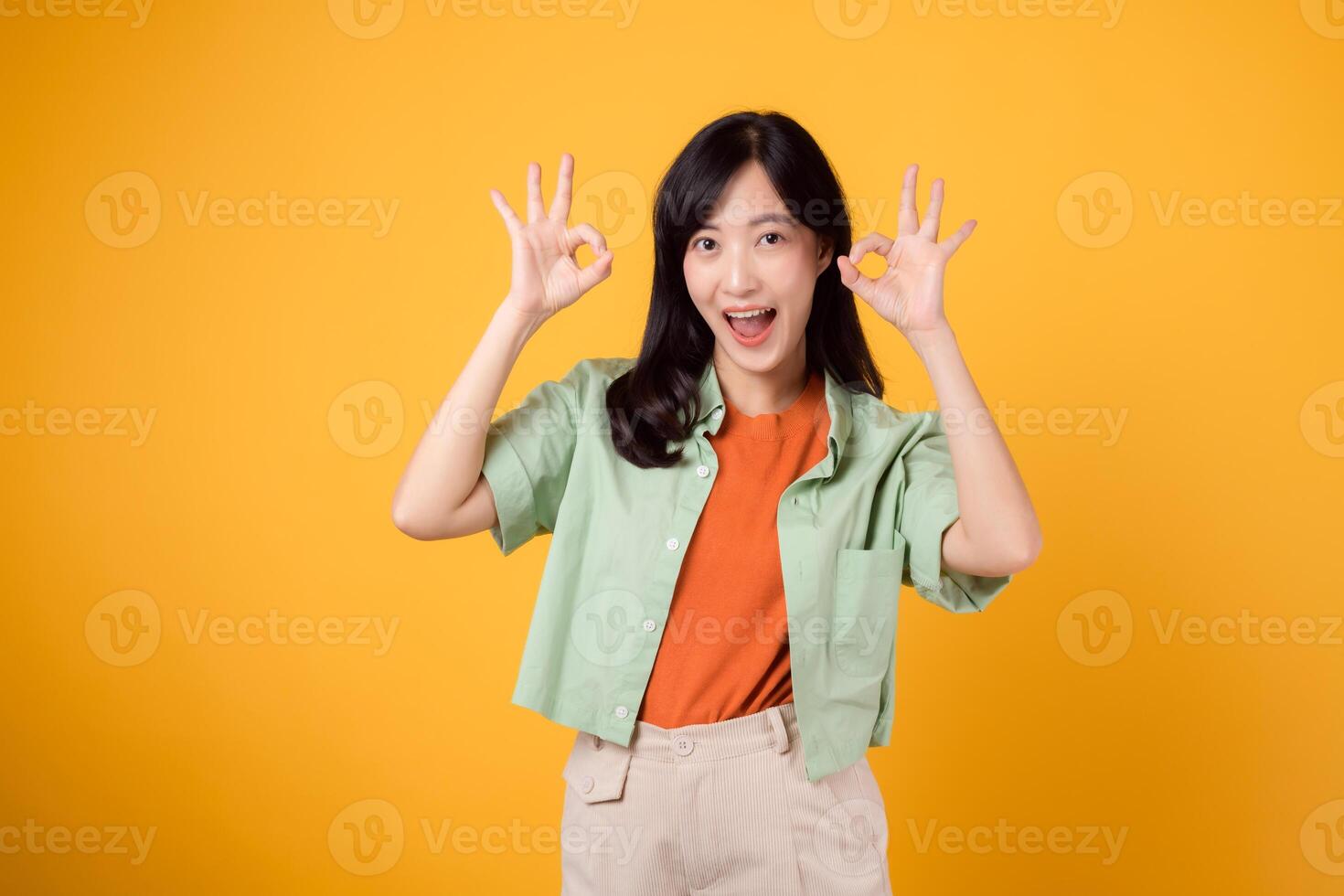joven asiático mujer 30, esmeradamente vestido en naranja camisa y verde puente. su bueno mano gesto y amable sonrisa, capturado en amarillo fondo, transmitir un reconfortante mensaje mediante cuerpo idioma. foto