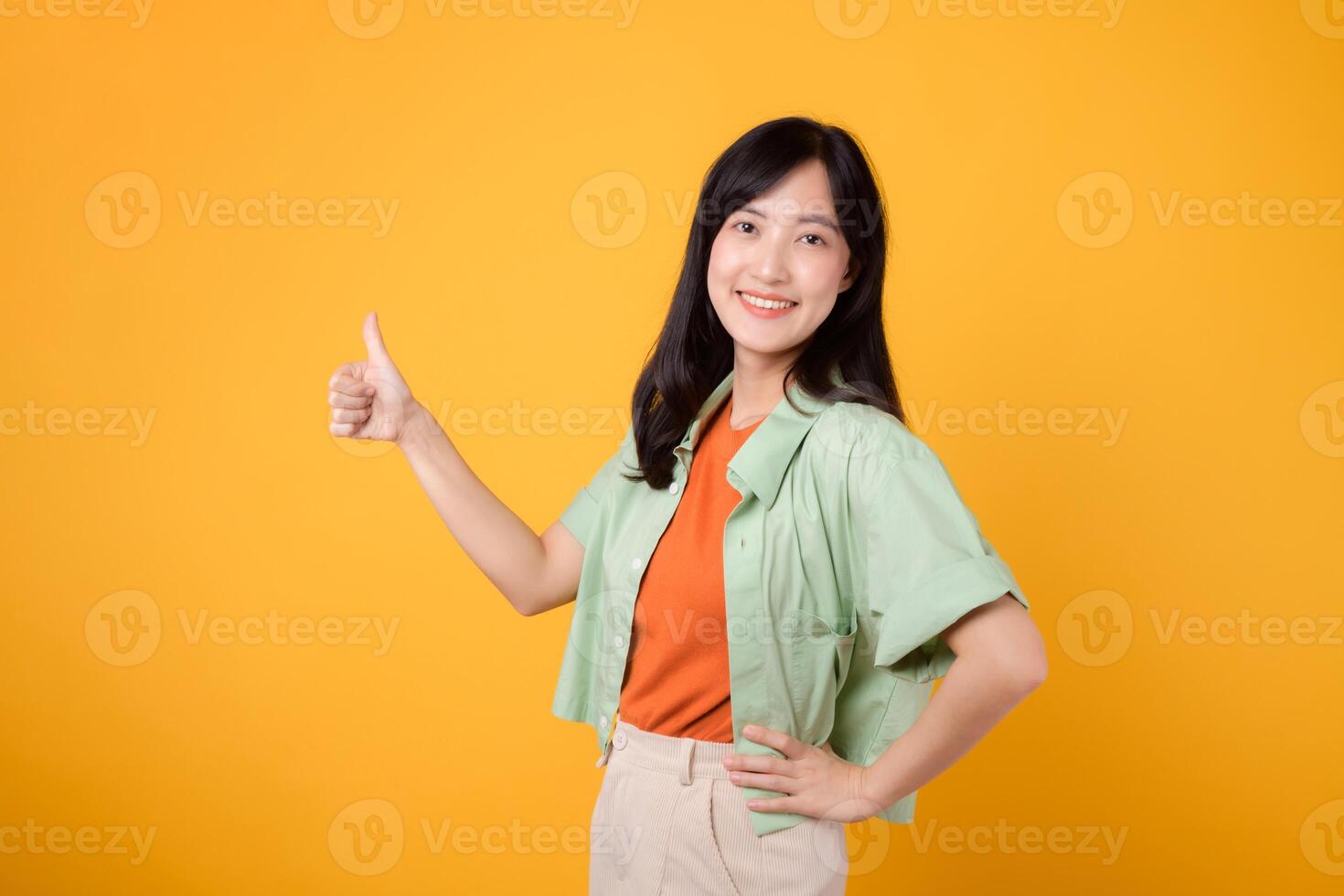 aprobación con alegre joven asiático mujer 30, esmeradamente vestido en naranja camisa y verde puente. su pulgares arriba gesto, conjunto en contra vibrante amarillo fondo, refleja el concepto de ánimo. foto