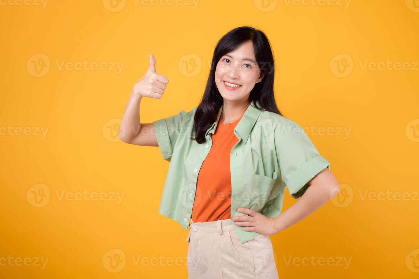 alegre joven asiático mujer en su 30, esmeradamente vestido en un naranja camisa y verde puente. su pulgares arriba gesto, conjunto en contra un vibrante amarillo fondo, refleja el concepto de ánimo. foto