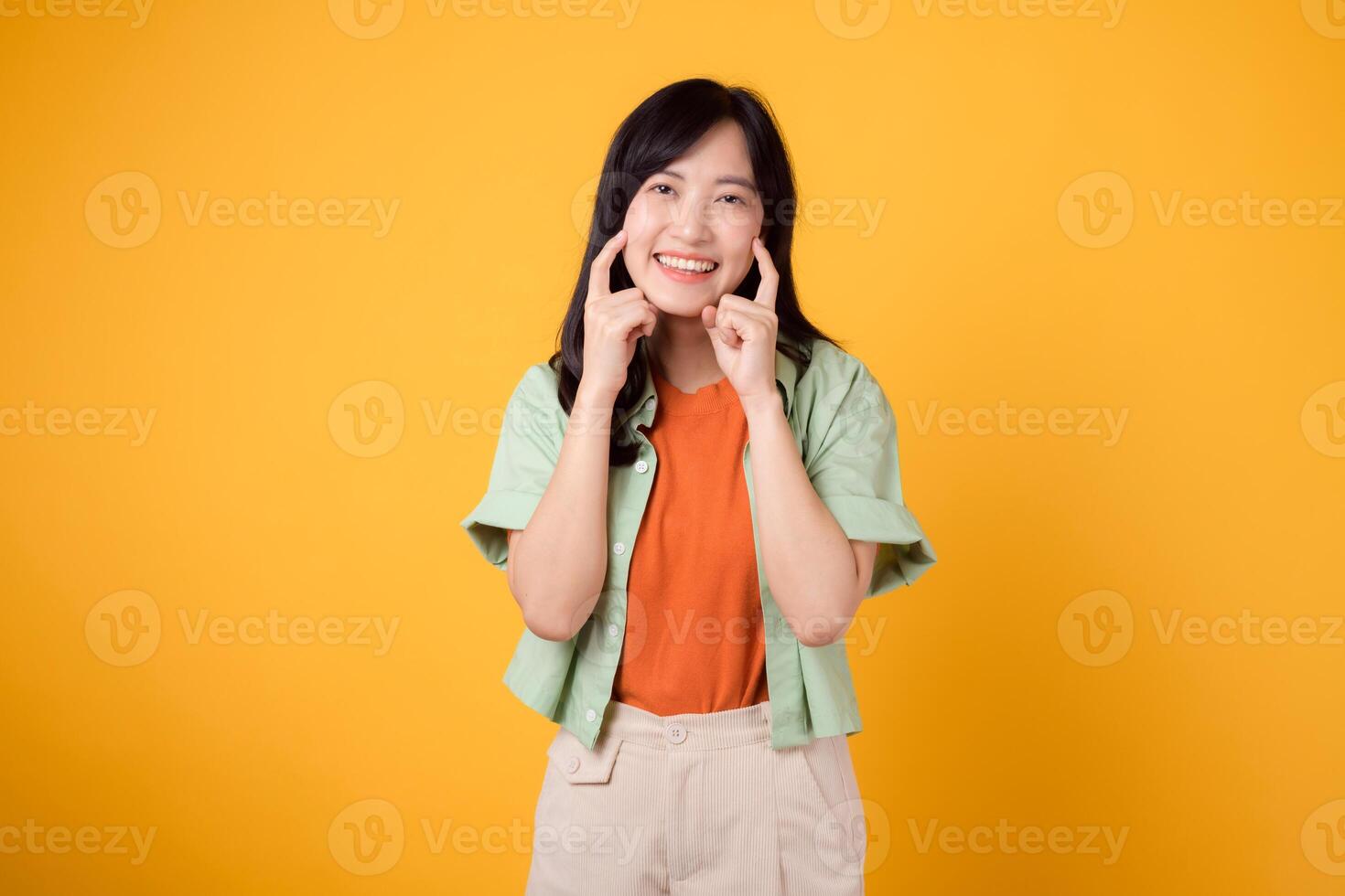 dental higiene con encantador joven asiático mujer 30, esmeradamente revestido en naranja camisa y verde saltador puntos a su dientes en un amarillo fondo, destacando el significado de dental cuidado de la salud. foto