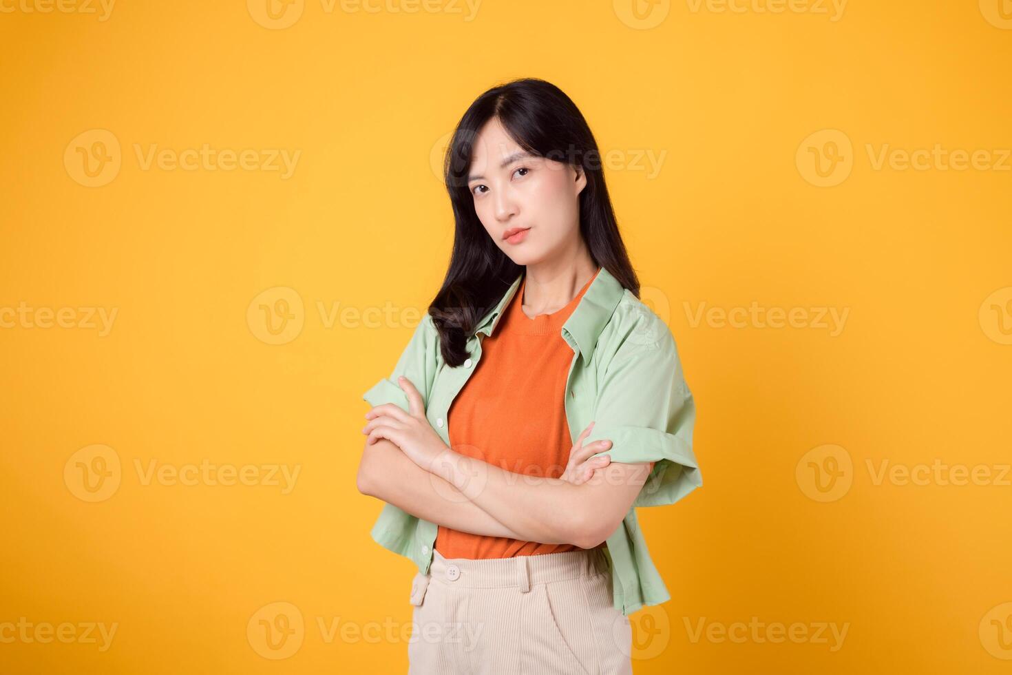 confianza y enojado con un joven asiático mujer 30s vistiendo naranja camisa. su brazo cruzar gesto en cofre en contra un vibrante amarillo antecedentes foto