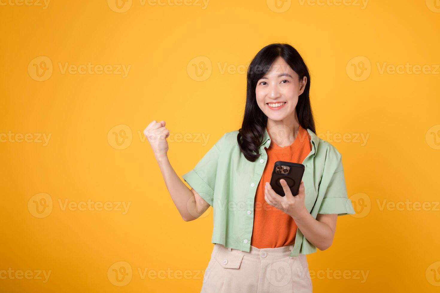 Excited young Asian woman in her 30s, wearing orange shirt and green jumper, using smartphone with fist up gesture on yellow studio background. Great news from mobile concept. photo