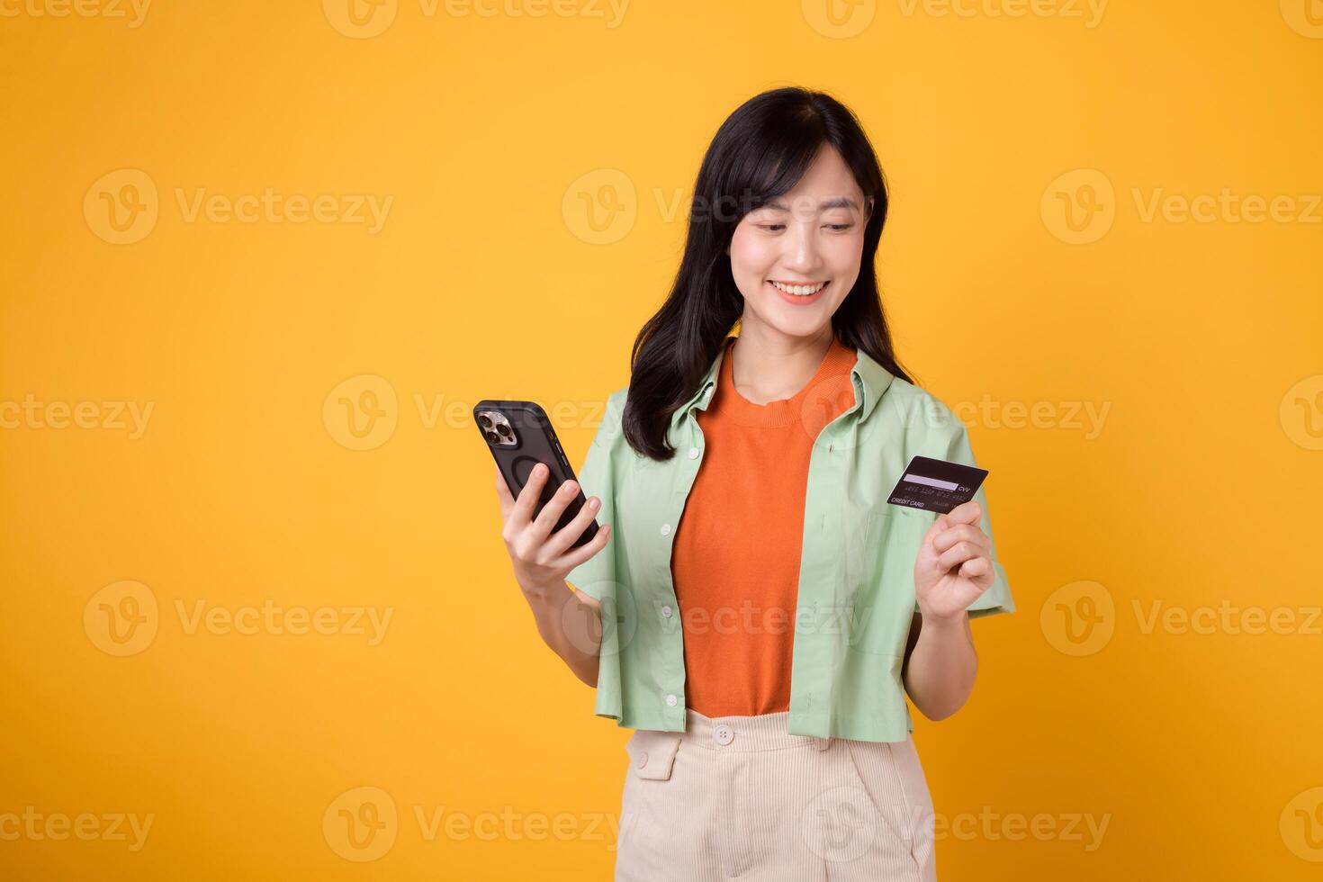 alegría de en línea compras con un alegre joven asiático mujer en su 30, vistiendo naranja camisa y verde saltador, utilizando teléfono inteligente mientras participación crédito tarjeta en amarillo estudio antecedentes. móvil concepto. foto