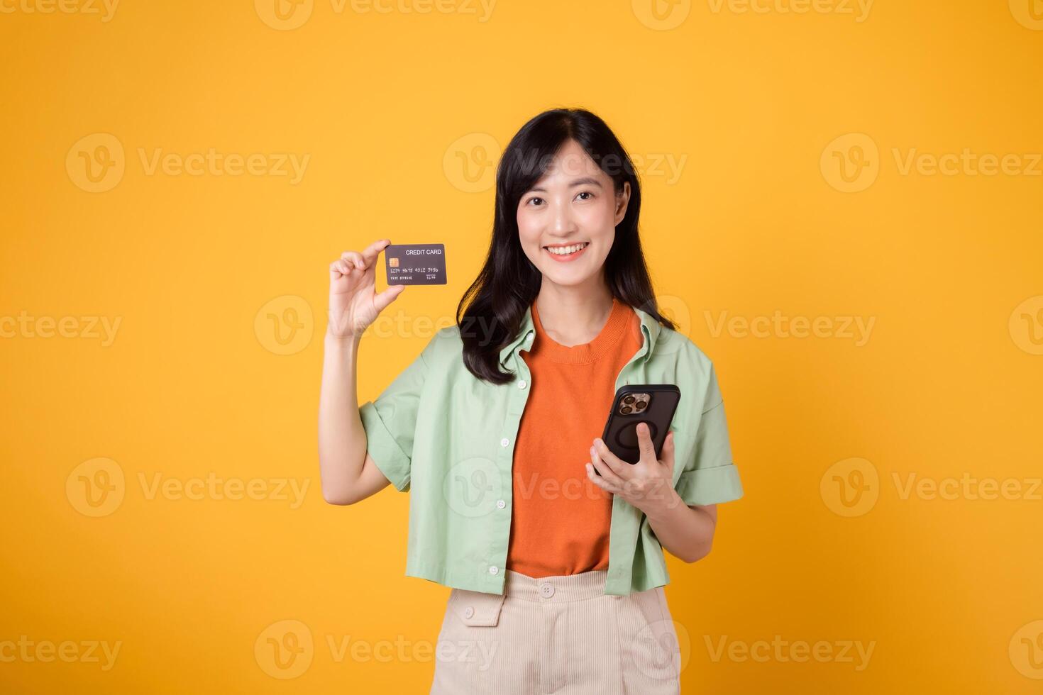abrazo el conveniencia de móvil compras con un encantador joven asiático mujer en su 30, vistiendo naranja camisa y verde saltador, utilizando teléfono inteligente a espectáculo crédito tarjeta en amarillo estudio antecedentes. foto