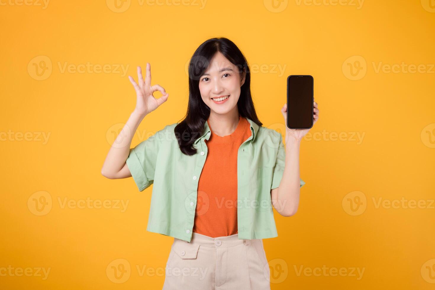 emoción de un nuevo móvil solicitud con un alegre joven asiático mujer 30, poniéndose naranja camisa y verde saltador, mostrando teléfono inteligente pantalla con bueno mano gesto en amarillo estudio antecedentes. foto