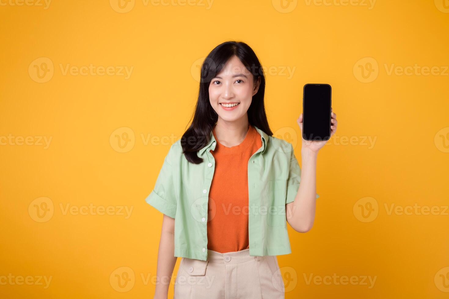 joven asiático mujer en su 30, vistiendo naranja camisa y verde saltador, vitrinas teléfono inteligente pantalla monitor en amarillo estudio antecedentes. nuevo móvil solicitud concepto. foto