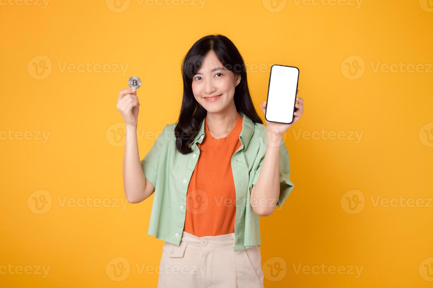joven 30s asiático mujer contento cara vestido en naranja camisa y verde saltador demostración teléfono inteligente pantalla monitor y cripto moneda moneda aislado en amarillo antecedentes. futuro Finanzas concepto. foto
