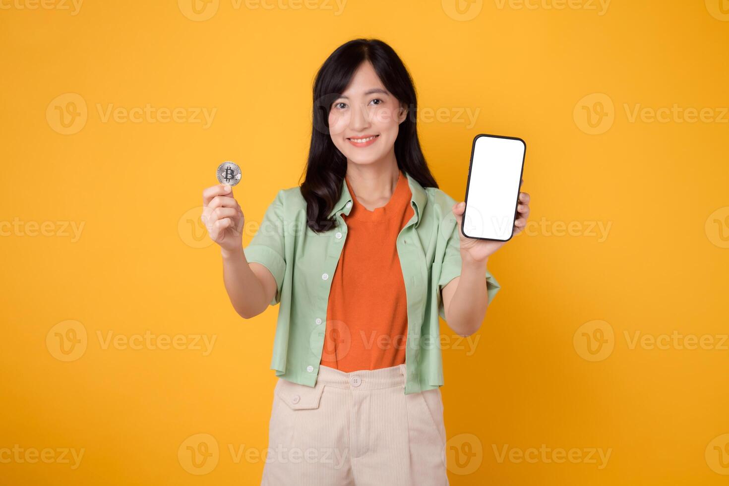 futuro Finanzas con un vibrante joven asiático mujer en su 30, vestido en naranja camisa y verde saltador, mostrando teléfono inteligente pantalla monitor y cripto moneda moneda en amarillo antecedentes. foto