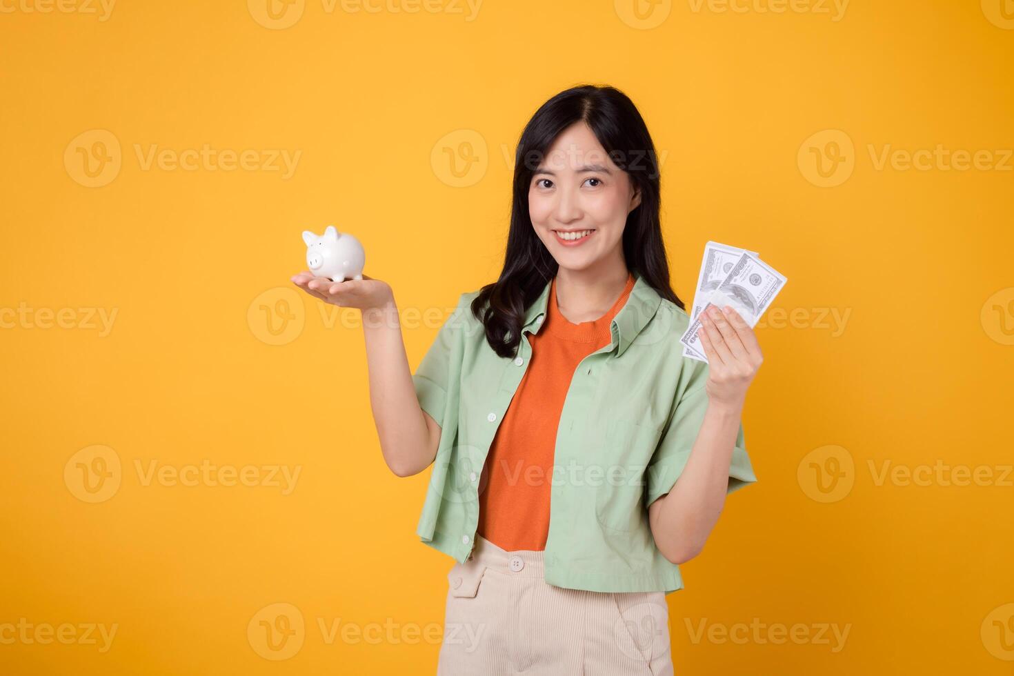 Happy young Asian woman in her 30s, wearing orange shirt and green jumper, showcases dollar currency and piggy bank on yellow background. Financial money concept. photo