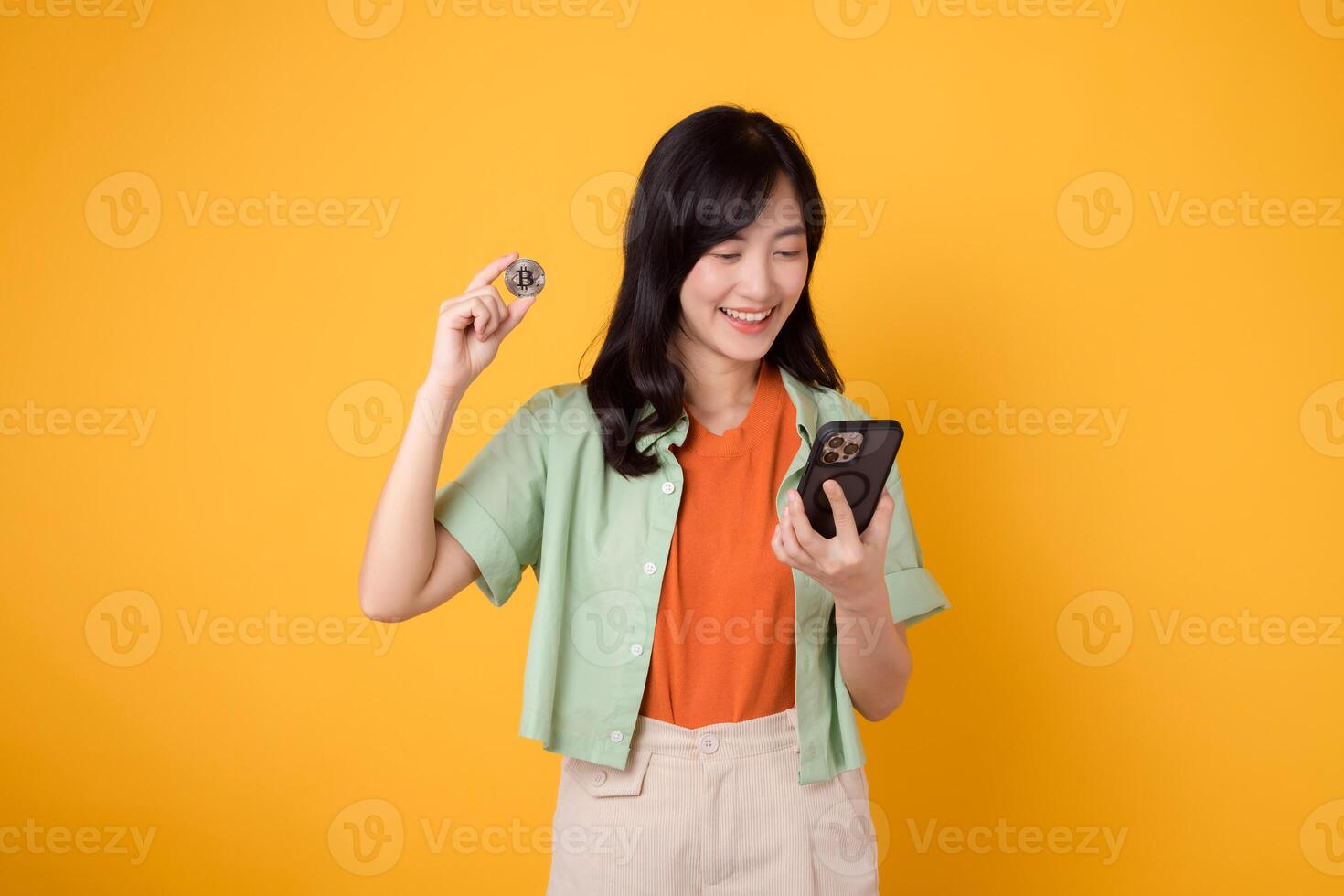 Embrace the future of finance with young Asian woman 30s, wearing orange shirt and green jumper, revealing crypto currency coin while using smartphone on yellow background. Future finance concept. photo