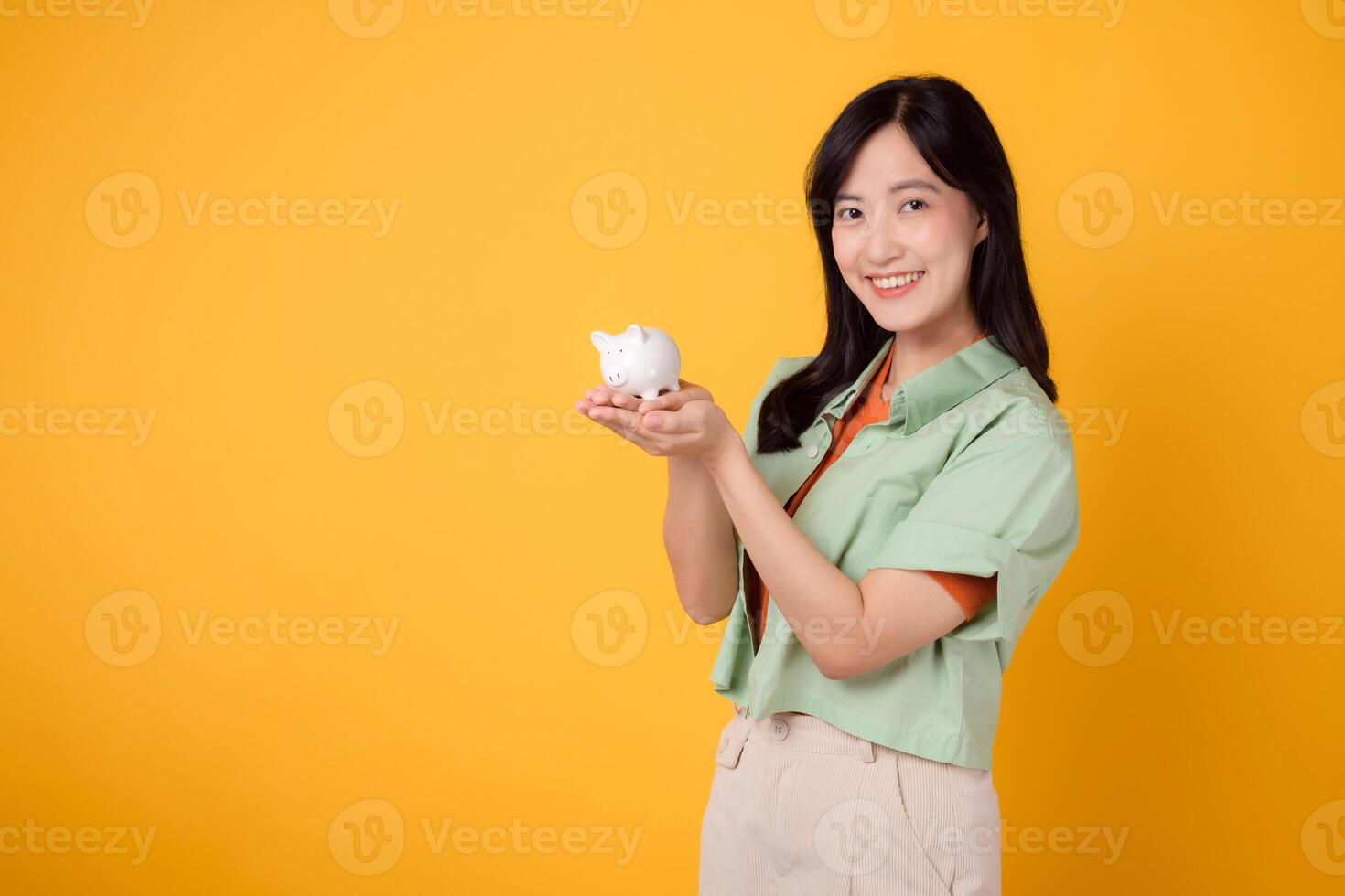 contento joven asiático mujer en su 30, vistiendo naranja camisa y verde saltador, vitrinas cerdito banco en amarillo antecedentes. financiero dinero concepto. foto