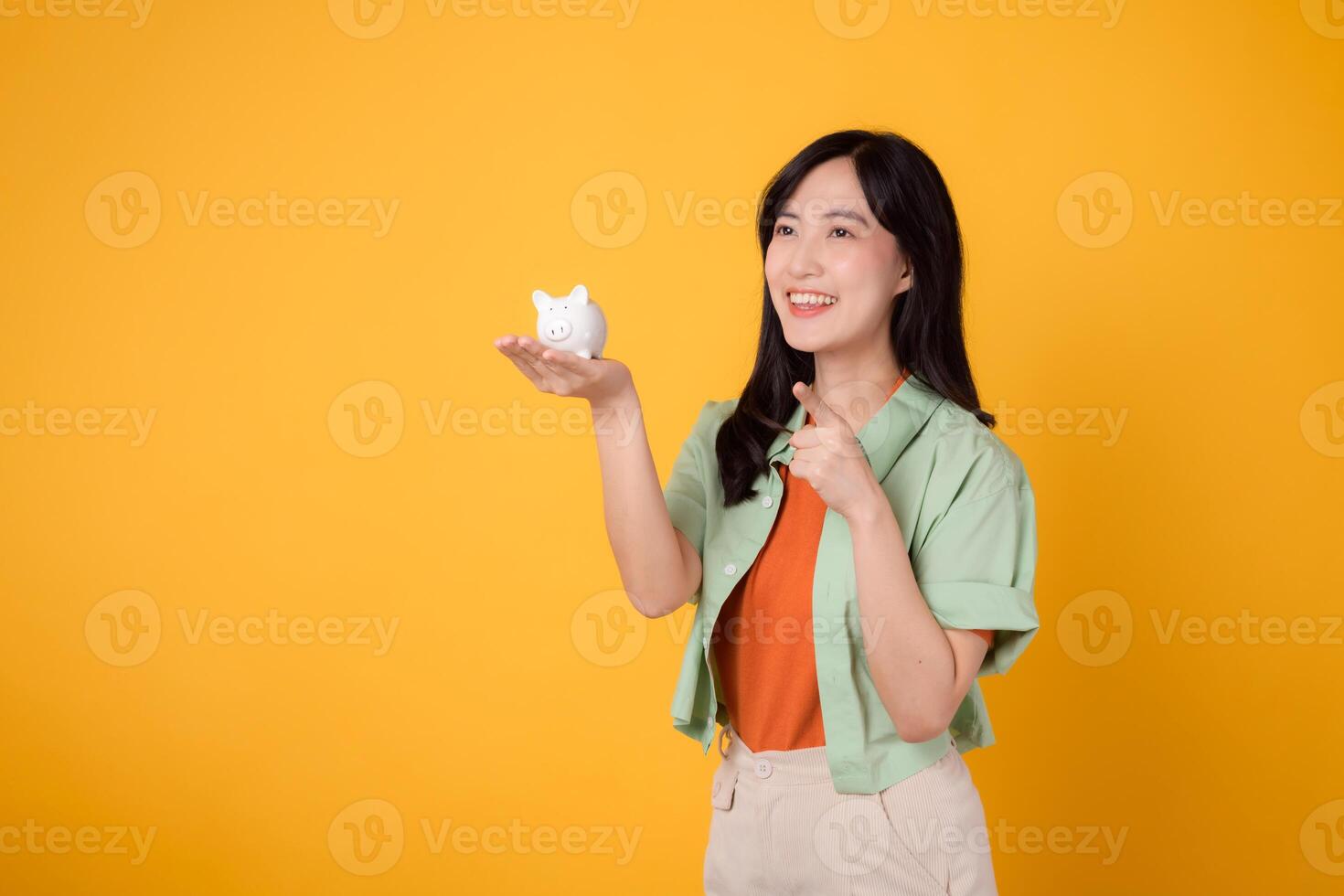 Happy young Asian woman in her 30s, wearing orange shirt and green jumper, showcases piggy bank while pointing finger to free copy space on yellow background. Financial money concept. photo