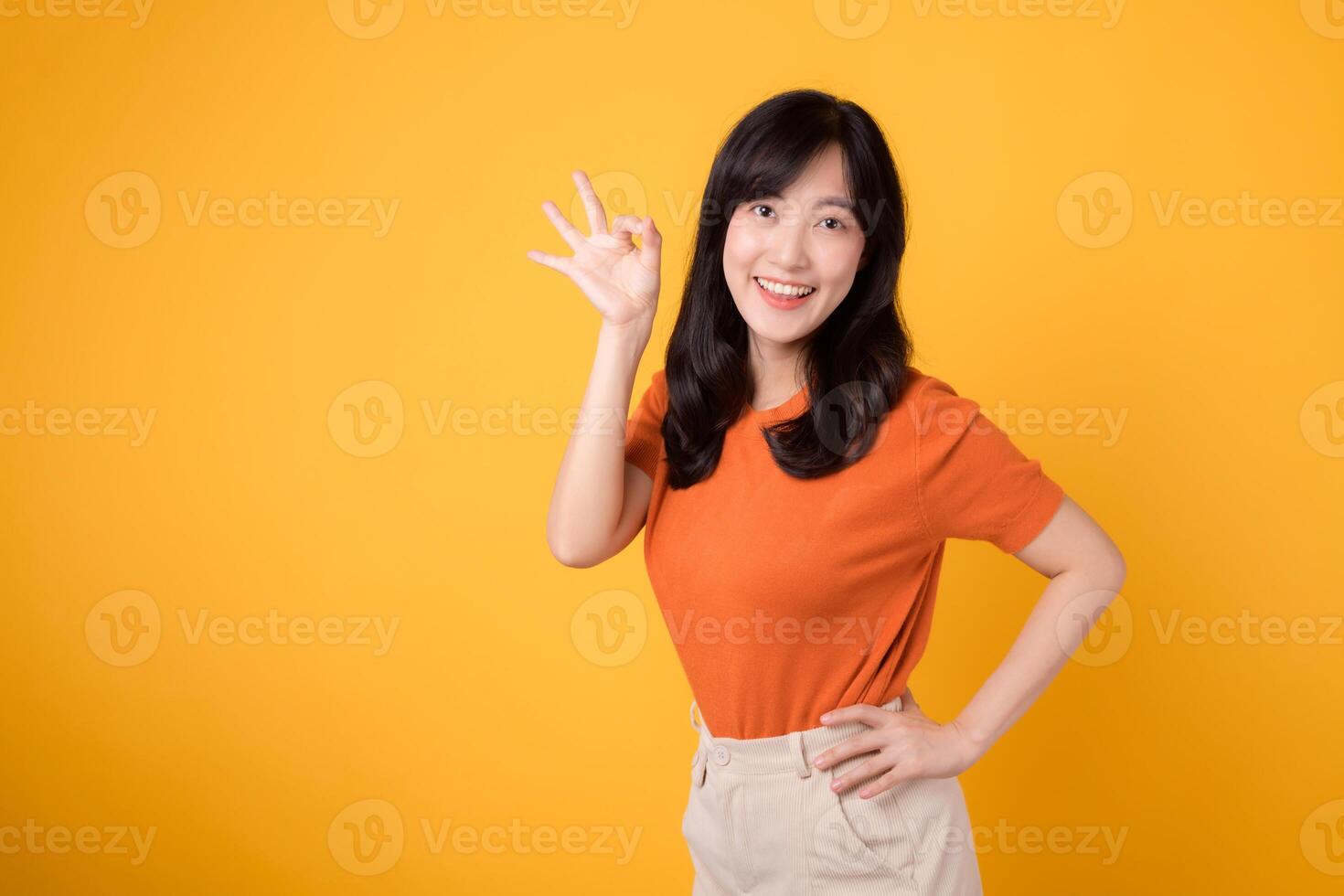 Embodying positivity, young Asian woman 30s flaunts okay sign in orange shirt on yellow background. Hand gesture concept. photo