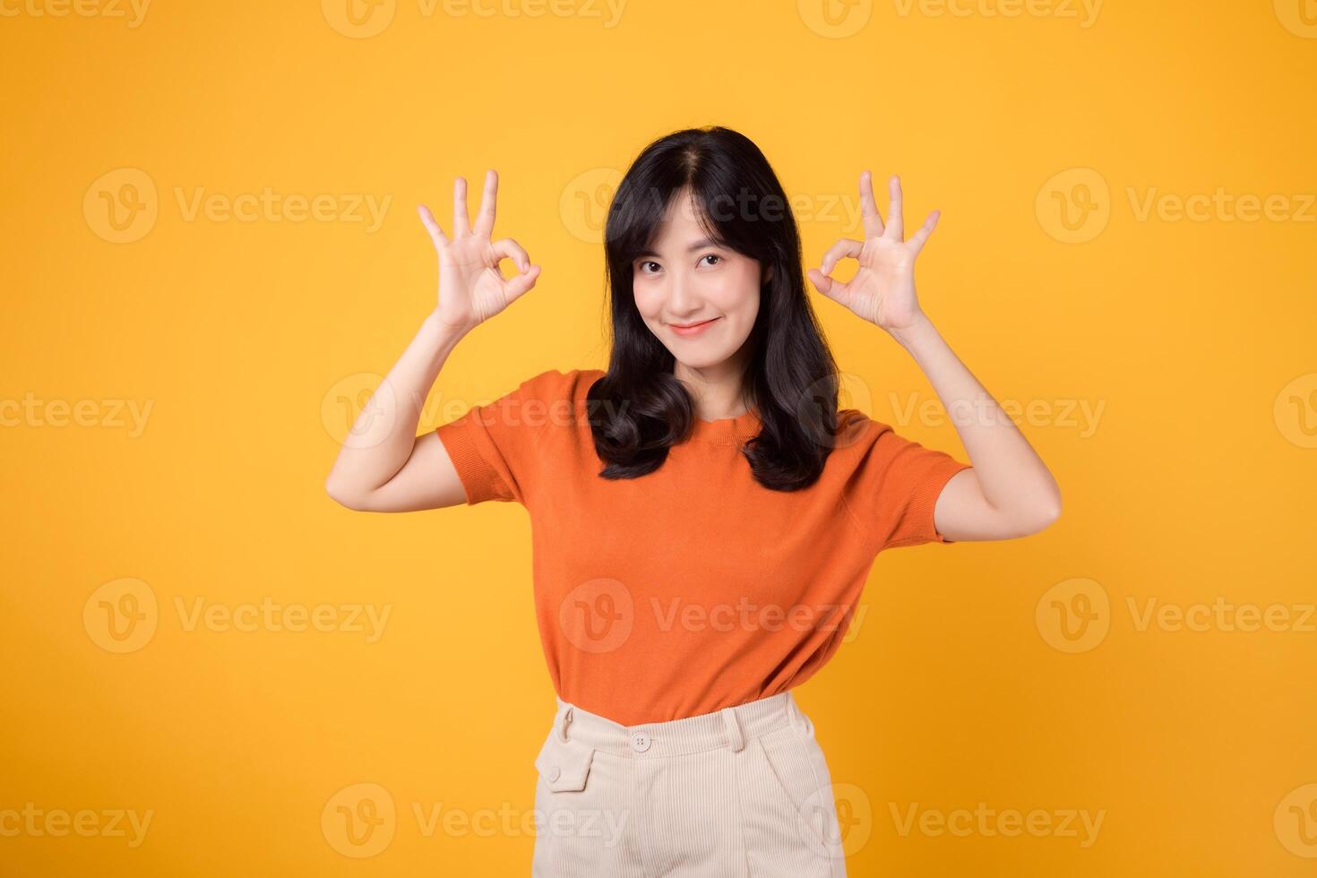 A confident young Asian woman in her 30s, dressed in an orange shirt, displays the okay sign gesture on a bright yellow background. Hands gesture concept. photo