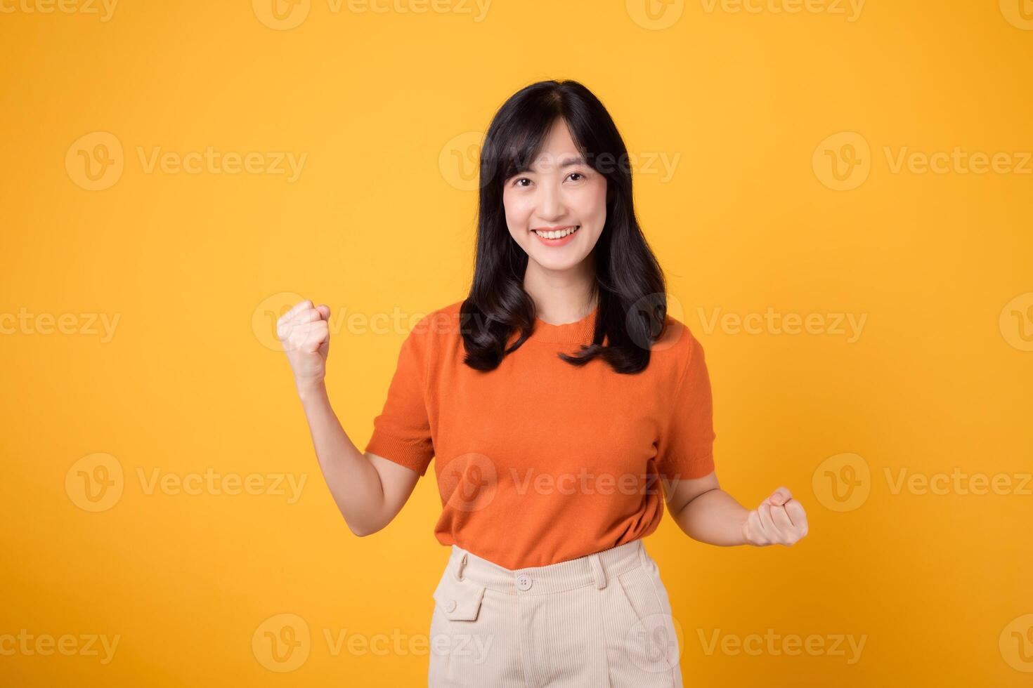 Embrace confidence as a young Asian woman in her 30s raises a fist up hand sign gesture, wearing an orange shirt on yellow background. photo
