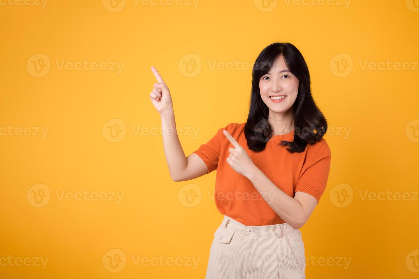 Radiate happiness with a young Asian woman in her 30s, dressed in an orange shirt, pointing to free copy space on yellow background. recommending product concept. photo