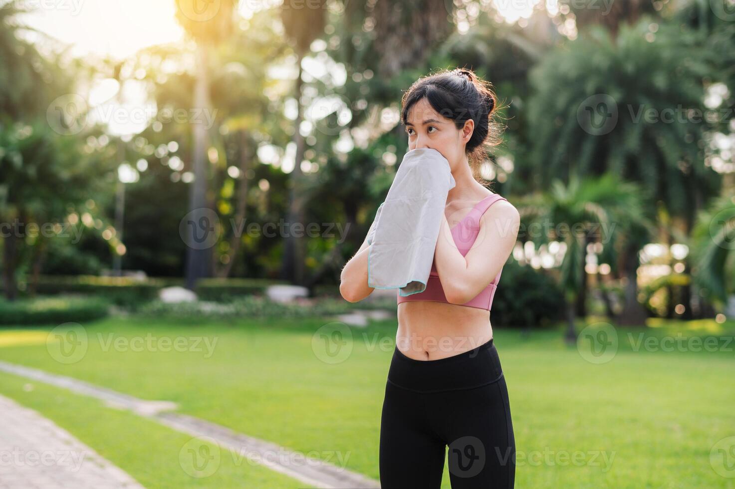 beauty of a healthy lifestyle as a 30s Asian woman in pink sportswear wipes her face after a sunset run in the public park. Achieve your fitness goals and embrace a sense of accomplishment. photo