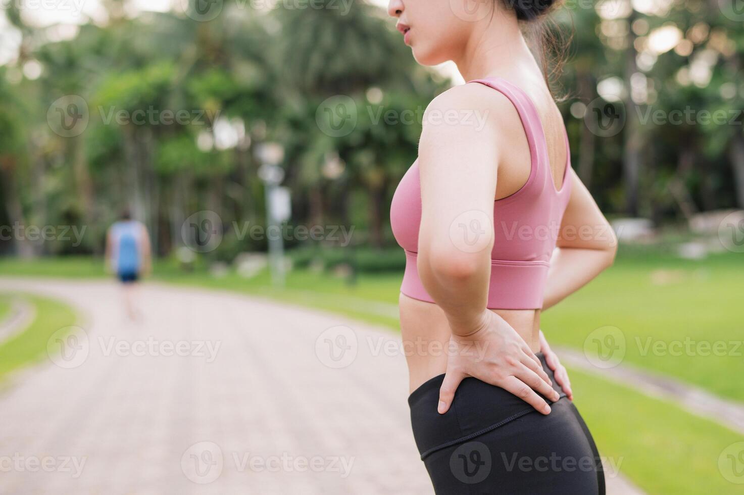 Woman jogger. young 30s asian female wearing pink sportswear holding her back and hip pain after running exercise in public park. Pain in activity concept. photo