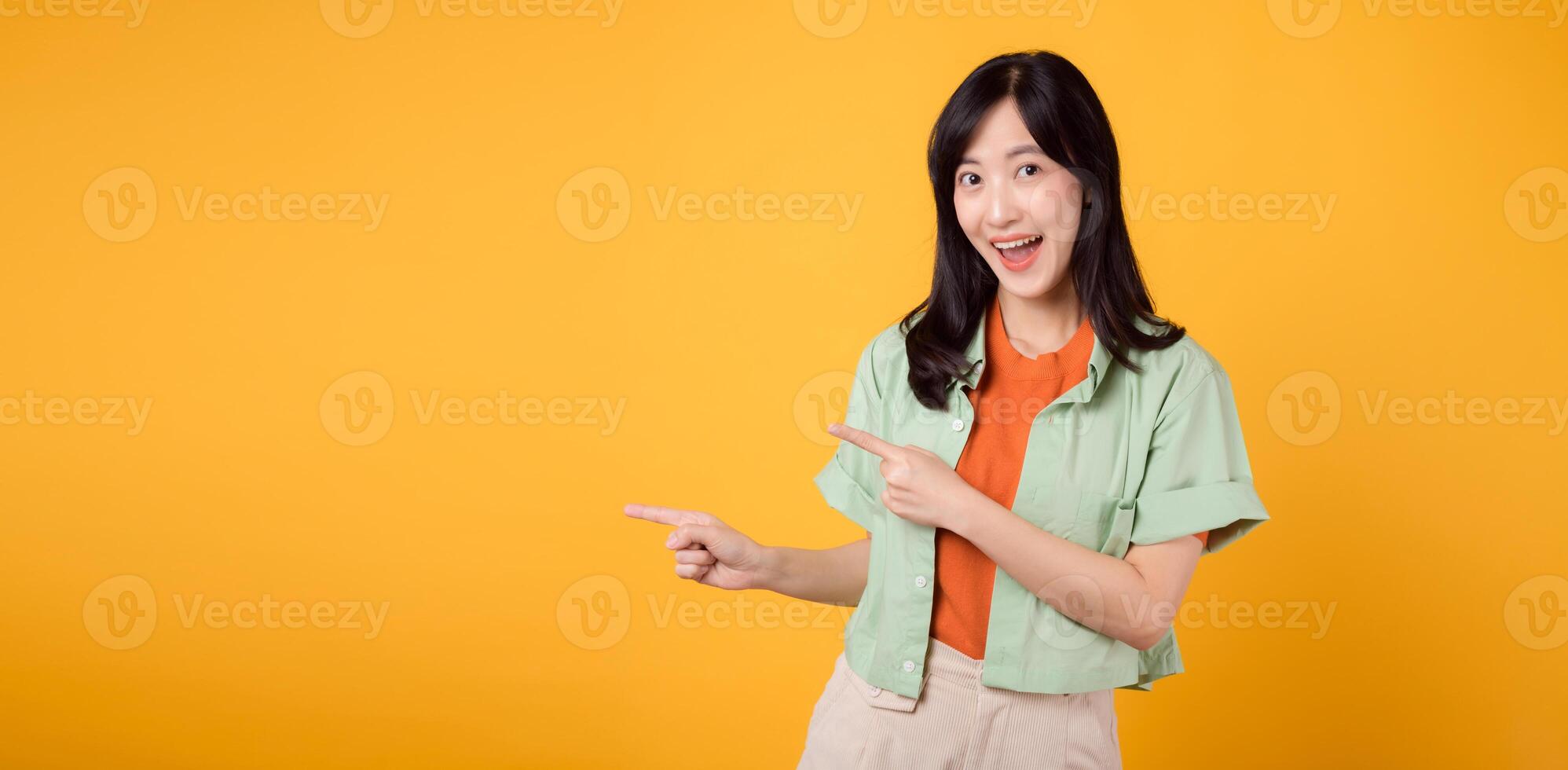 young Asian woman in her 30s, wearing a green shirt on an orange shirt, pointing fingers to free copy space. Explore the concept of discount shopping promotion with this eye-catching visual. photo