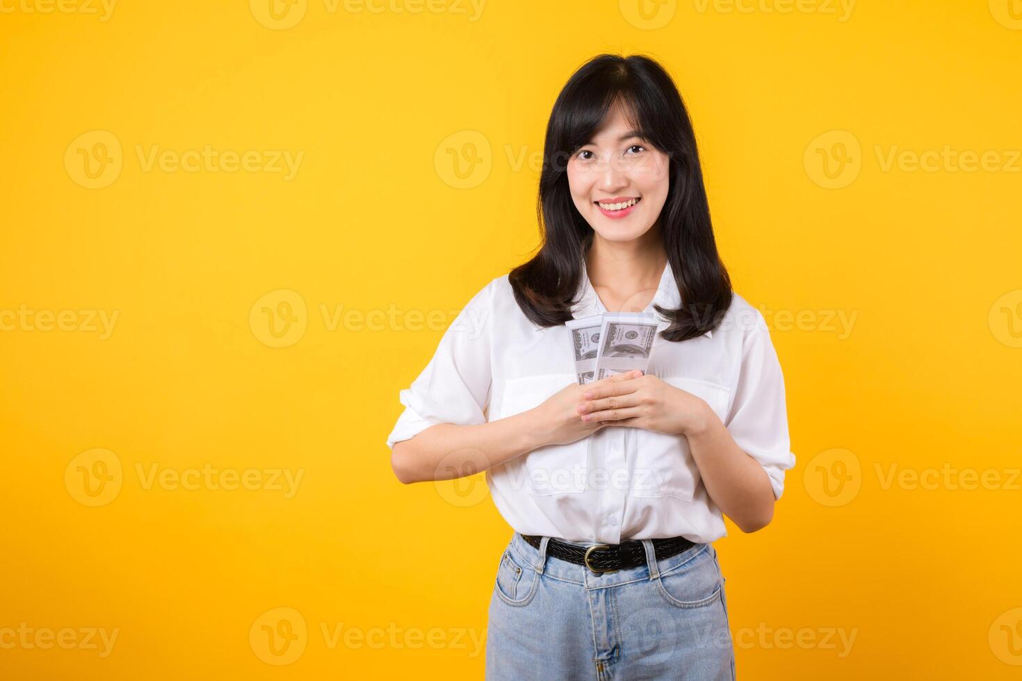 Happy young asian businesswoman wearing white shirt and denim jean holding cash, hugging dollars money and smiling, standing over yellow background. Love to be rich concept. photo