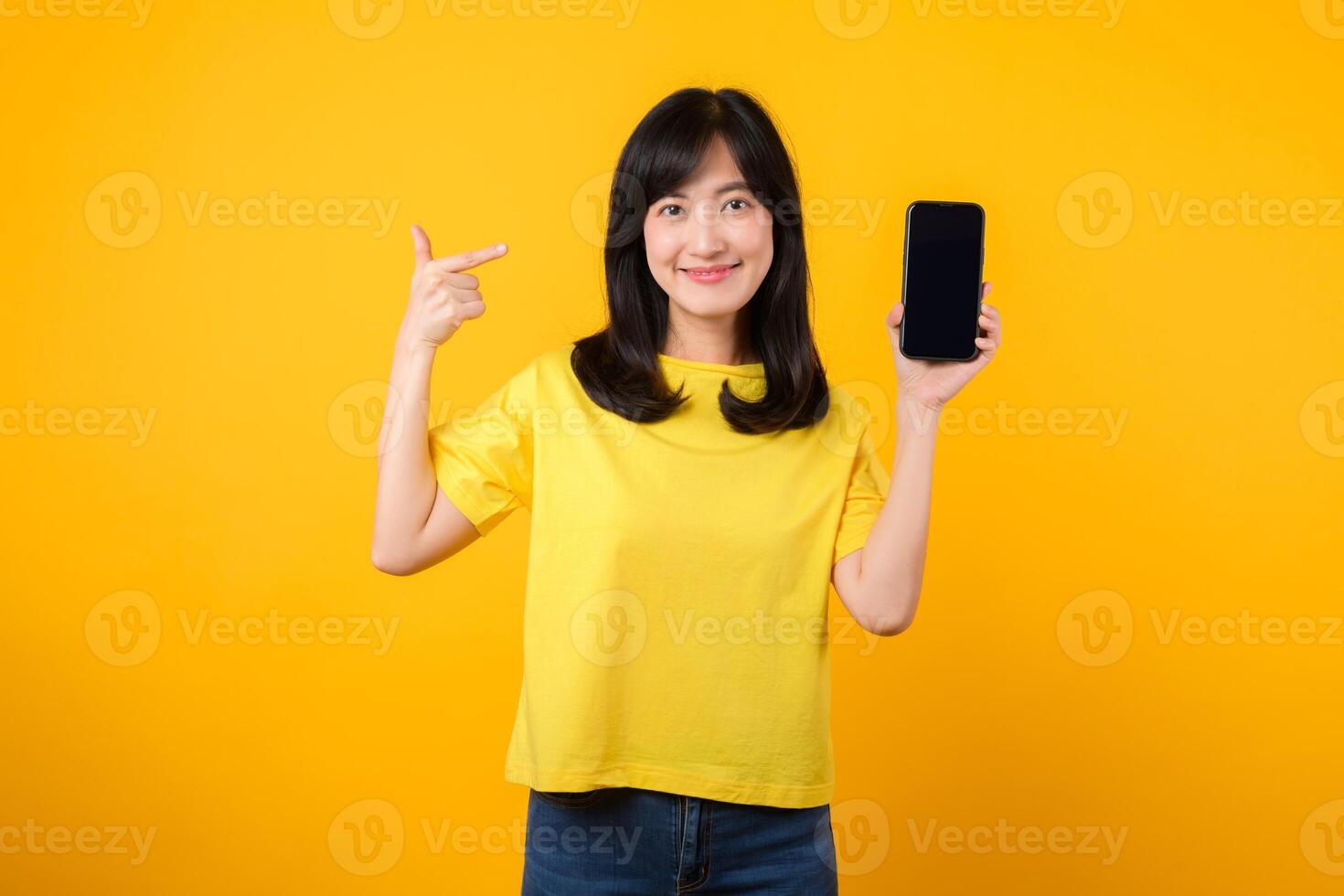 Capture joy and excitement of new applications with cheerful portrait young Asian woman wearing yellow t-shirt and denim jeans points finger towards smartphone. concept of app and digital innovation. photo