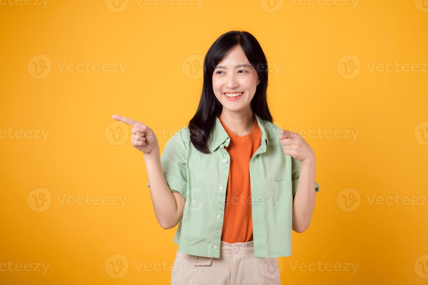 young Asian woman 30s wearing a green shirt on an orange shirt, confidently pointing fingers to free copy space. Explore the concept of discount shopping promotion with engaging image. photo