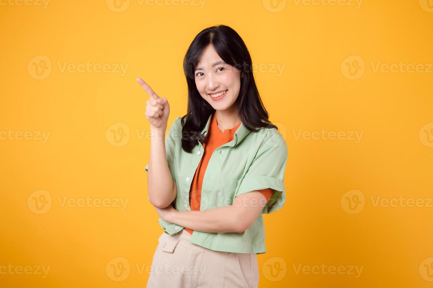 Unlock savings and shop now young Asian woman in her 30s, wearing a green shirt on an orange shirt. happy face and pointing finger to free copy space against vibrant yellow backdrop. photo