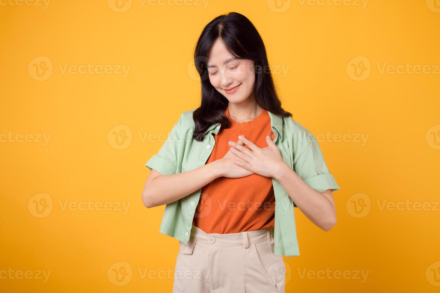 wellness as an Asian woman in her 30s, wearing a green shirt, holds her hand on her chest against an orange backdrop. Healthcare concept on a vibrant yellow background. photo