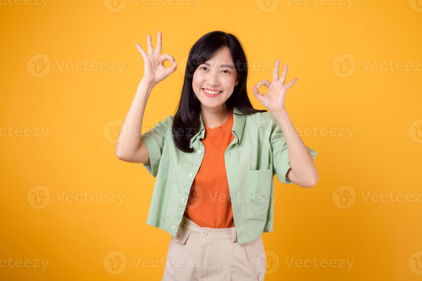 delightful young Asian woman 30s, dressed elegantly in orange shirt and green jumper. Her expressive okay hand gesture and gentle smile shine brightly on yellow background, body language concept. photo