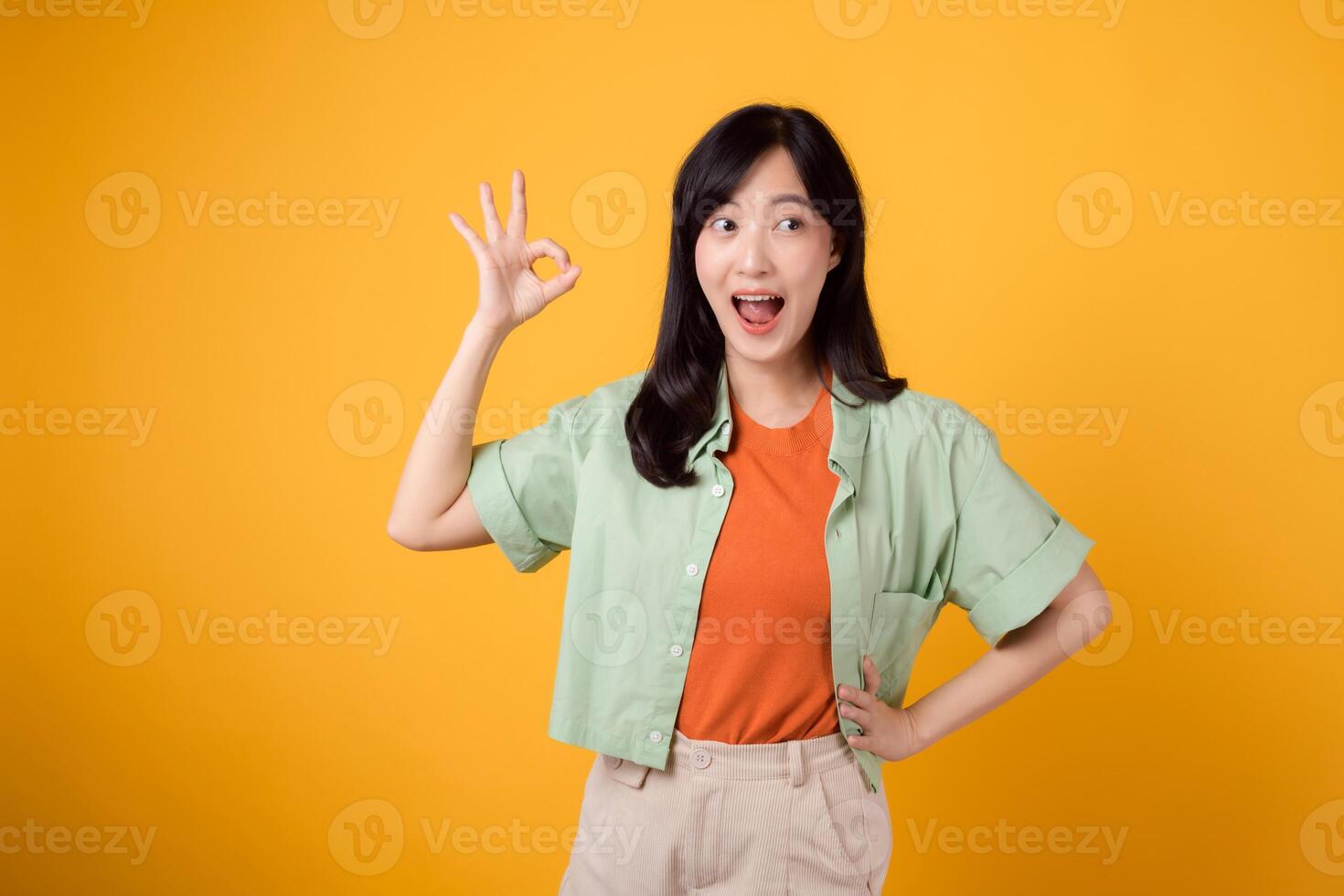 joven asiático mujer 30, esmeradamente revestido en naranja camisa y verde puente. su entrañable bueno mano gesto y amable sonrisa, conjunto en contra un amarillo fondo, revelar el belleza de cuerpo idioma. foto