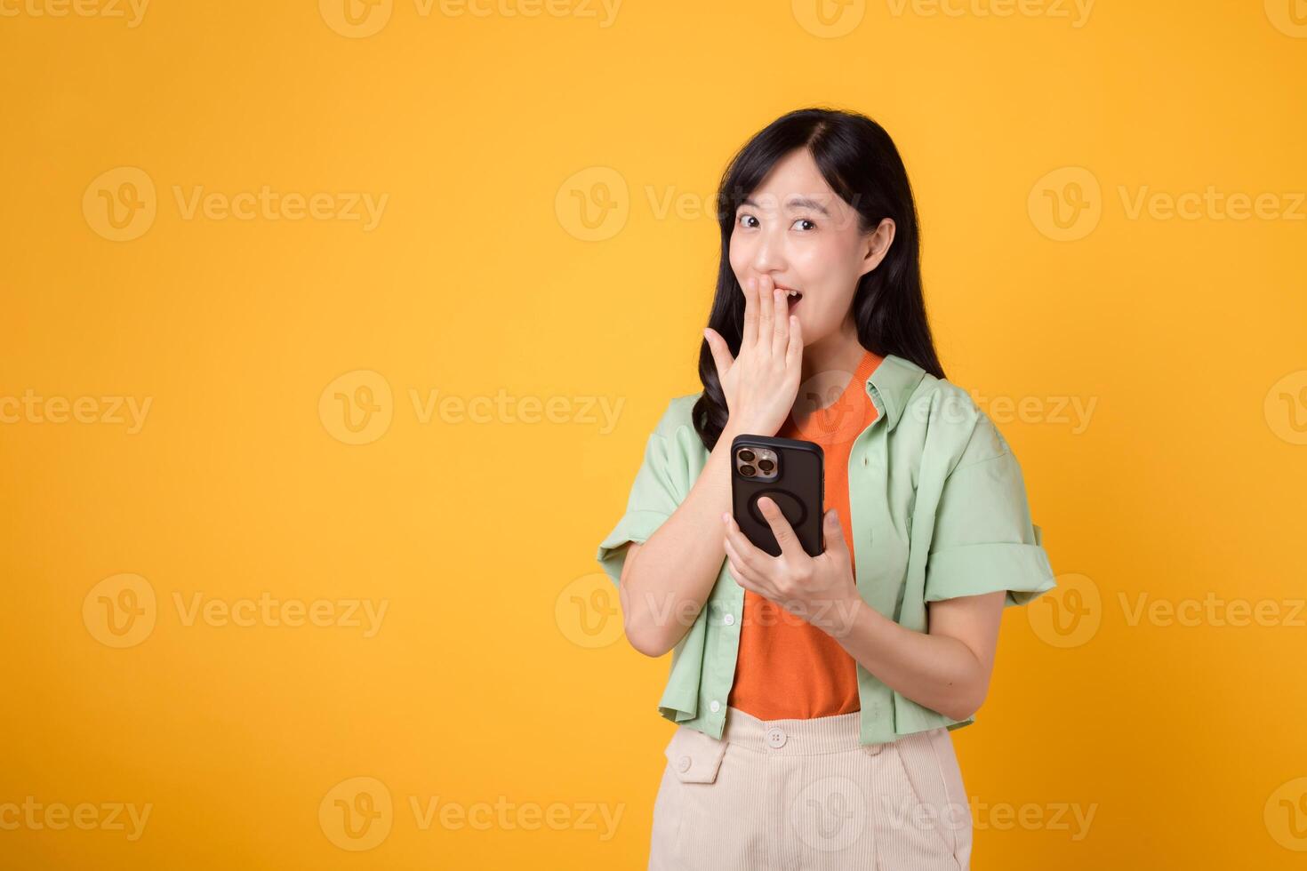 sorpresa como un joven asiático mujer en su 30, vistiendo un naranja camisa y verde saltador, es absorto en su teléfono inteligente en un amarillo estudio antecedentes. aplicación teléfono inteligente concepto. foto