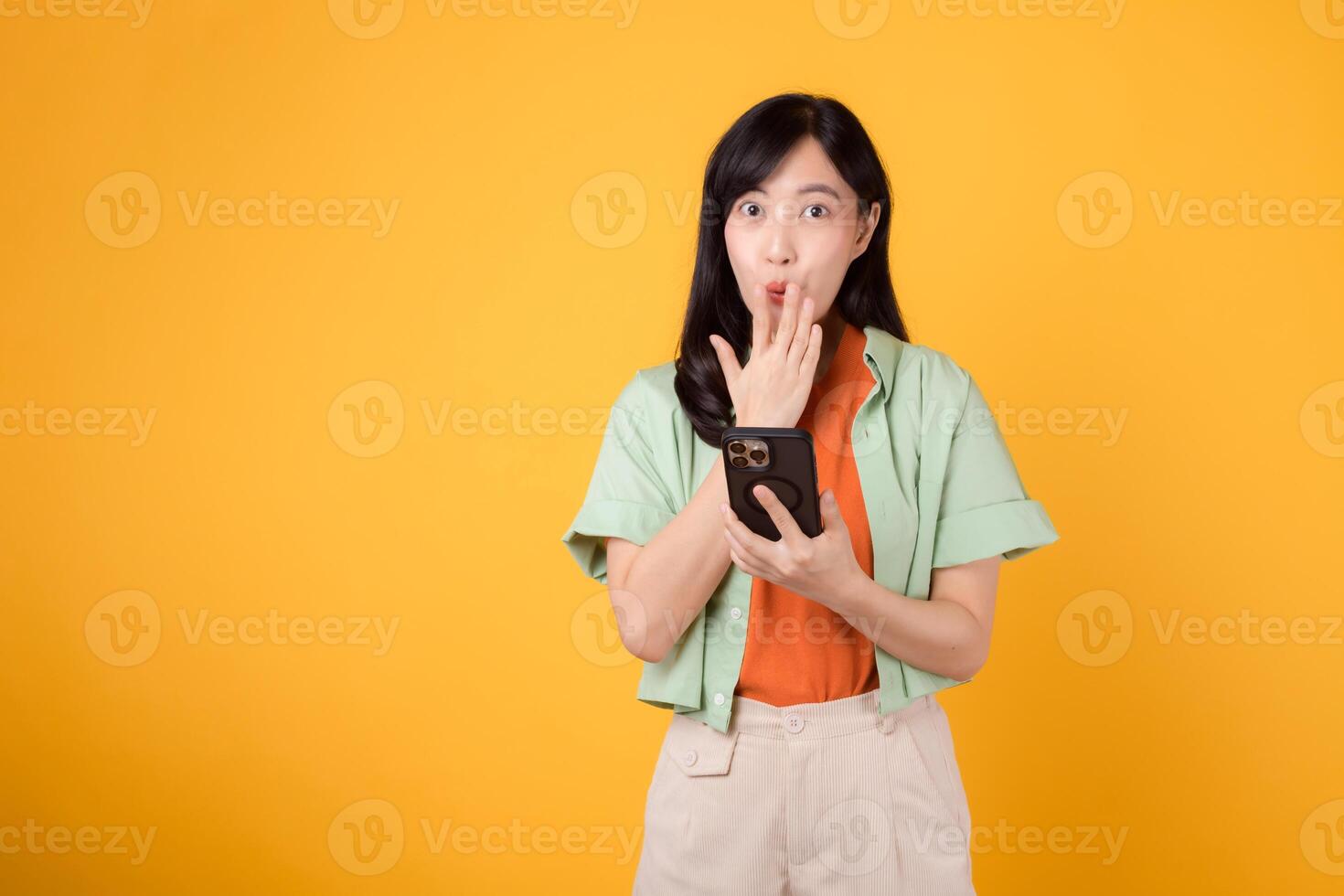 charm of surprise with a cheerful young Asian woman in her 30s, dressed in an orange shirt and green jumper, using her smartphone, mouth closed, on a yellow studio background. App smartphone concept. photo