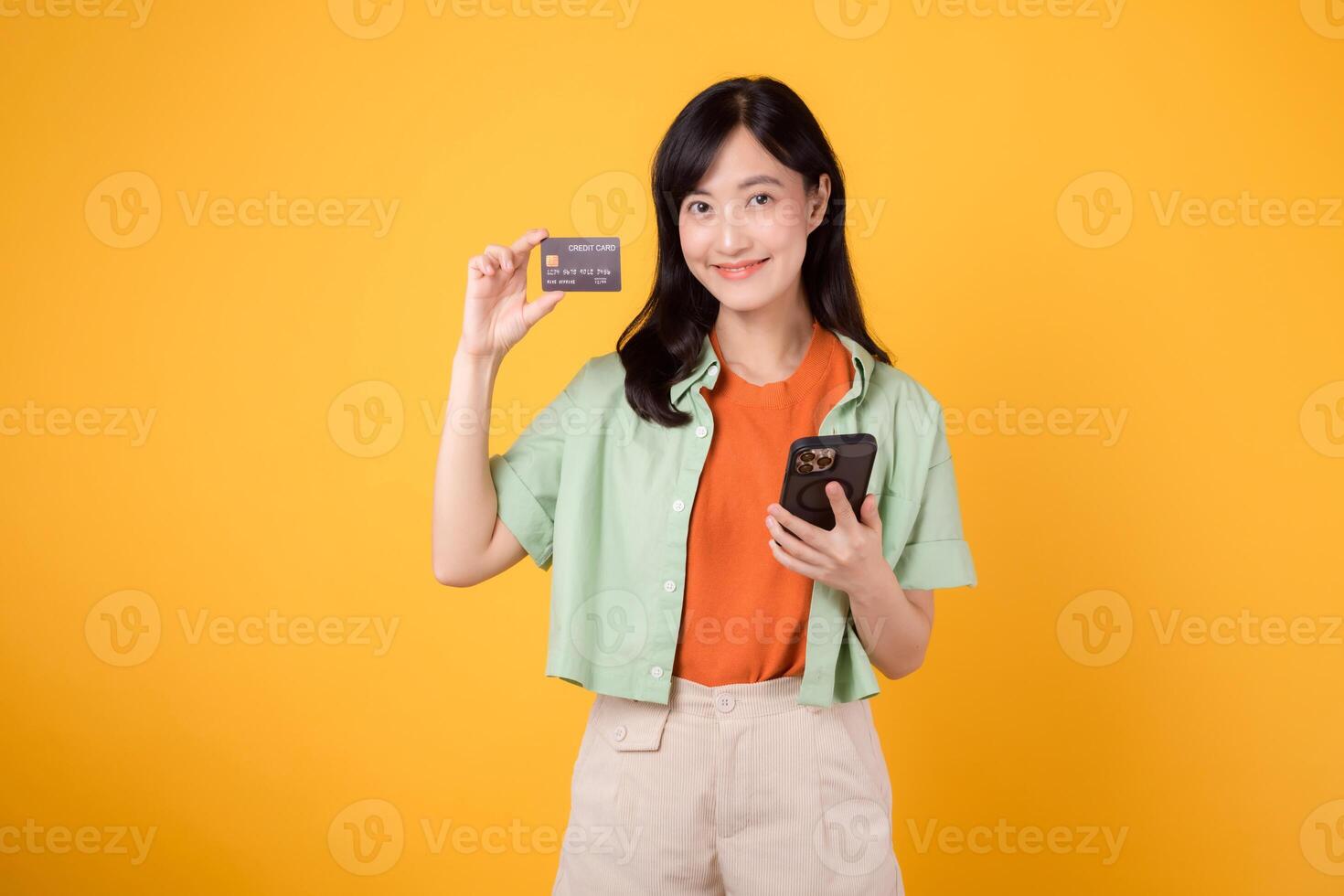 emocionado joven asiático mujer en su 30, vestido en naranja camisa y verde saltador, usos teléfono inteligente a espectáculo crédito tarjeta en amarillo estudio antecedentes. en línea compras desde móvil concepto. foto