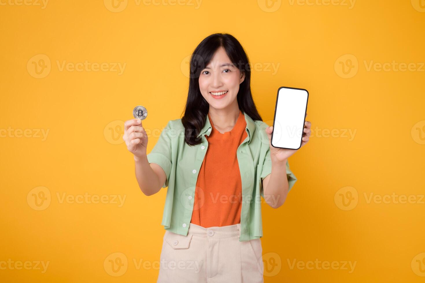 the innovation of future finance with young Asian woman in her 30s, elegantly dressed in orange shirt and green jumper, using smartphone screen display and crypto currency coin on yellow background. photo