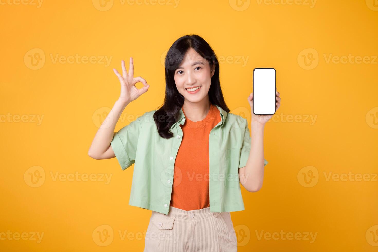 joven asiático mujer en su 30, vistiendo naranja camisa y verde saltador, vitrinas teléfono inteligente pantalla monitor con un bueno mano gesto en amarillo estudio antecedentes. nuevo móvil solicitud concepto. foto