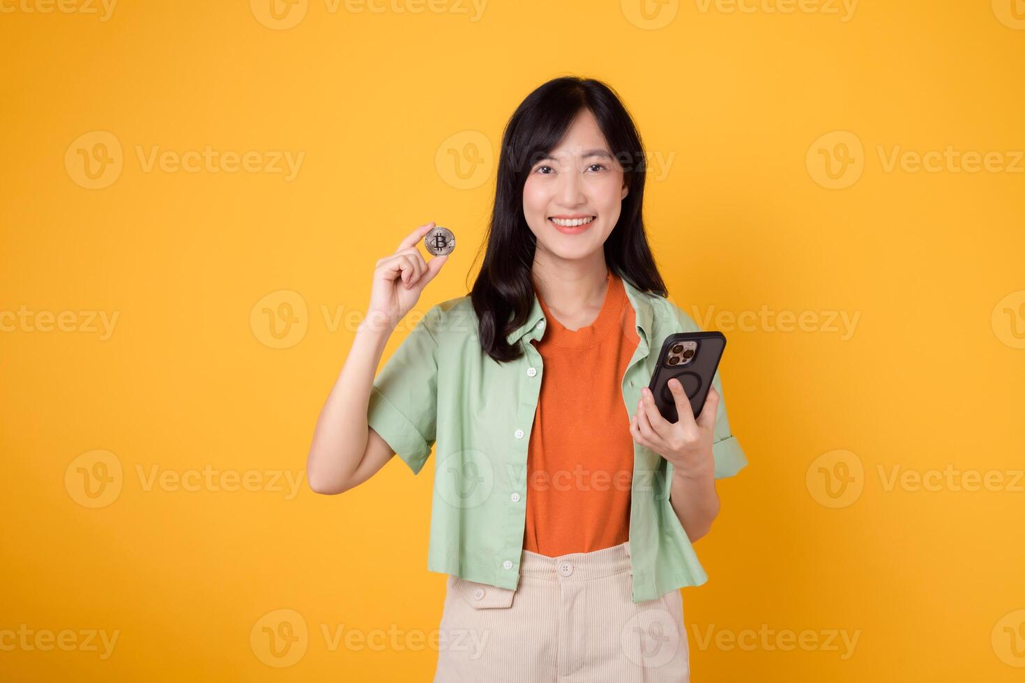 young Asian woman 30s, elegantly dressed in orange shirt and green jumper, showing crypto currency coin while holding smartphone on yellow background. Future finance concept. photo