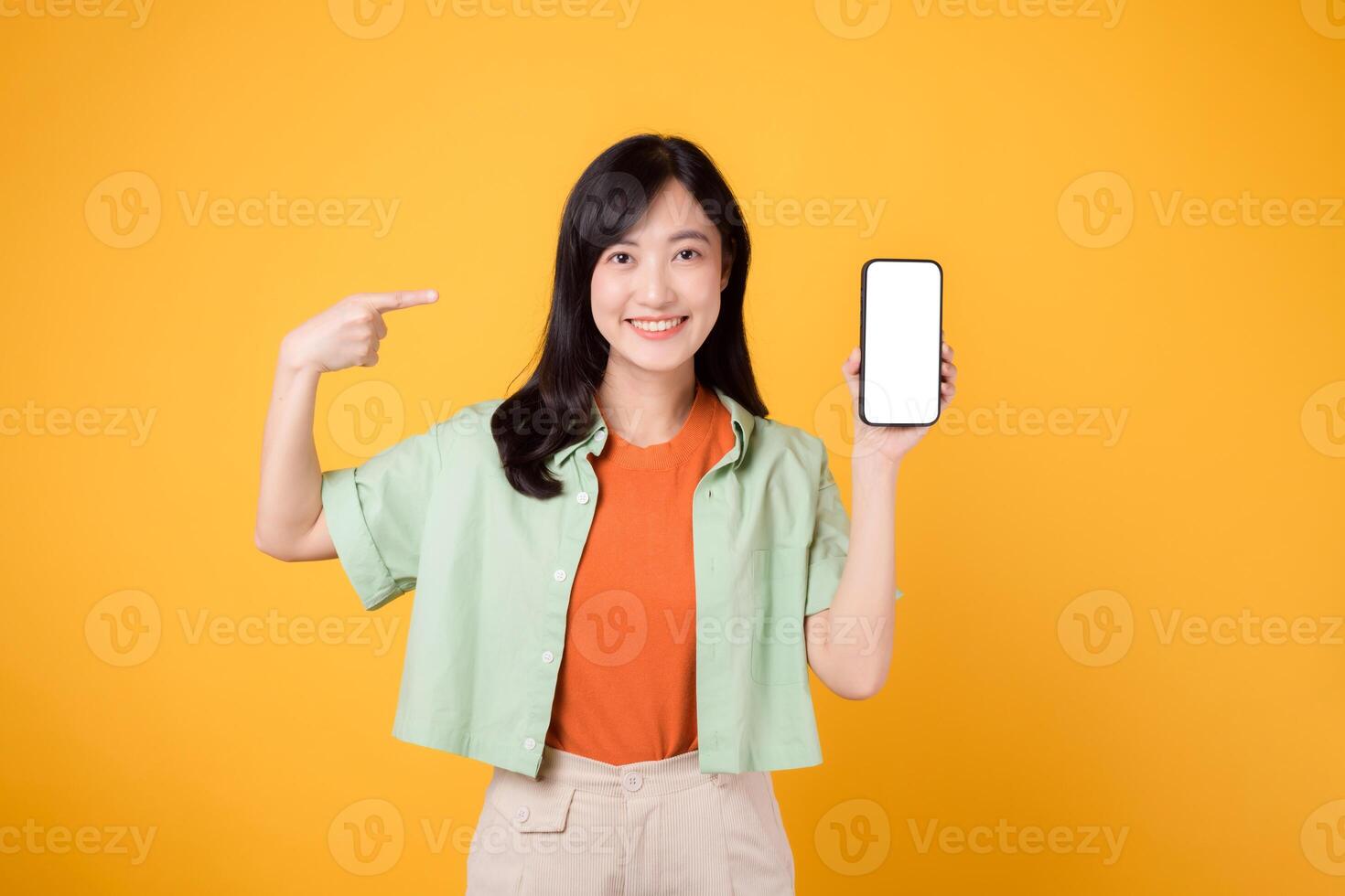 nuevo móvil solicitud con alegre joven asiático mujer en su 30, poniéndose naranja camisa y verde saltador, señalando dedo a mostrando teléfono inteligente pantalla en amarillo estudio antecedentes. foto