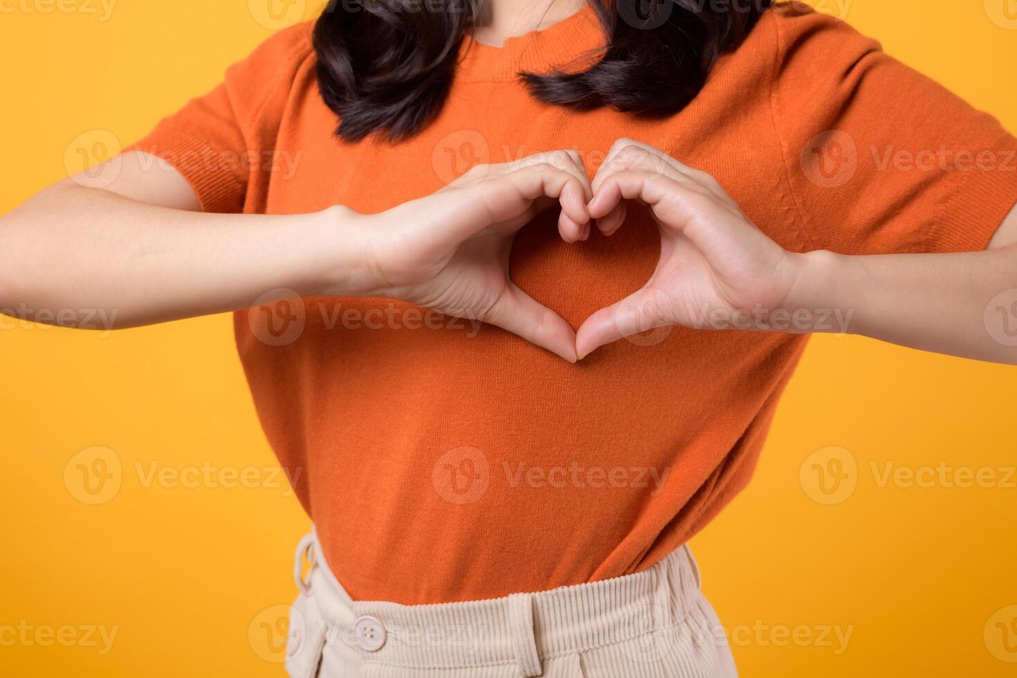 healthcare, wellness, and well-being with a young Asian woman 30s. She showcases a heart hand gesture on her chest, wearing an orange shirt, against a vibrant yellow background. photo