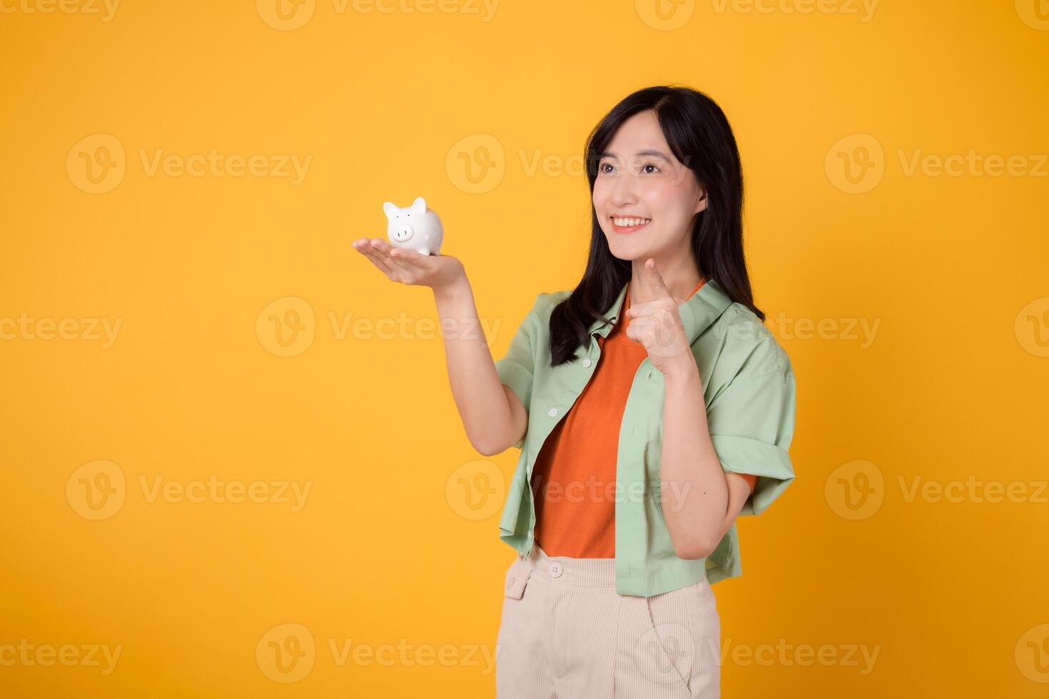 joven 30s asiático mujer contento cara vestido en naranja camisa y verde saltador demostración cerdito banco mientras señalando dedo a gratis Copiar espacio aislado en amarillo antecedentes. financiero dinero concepto. foto