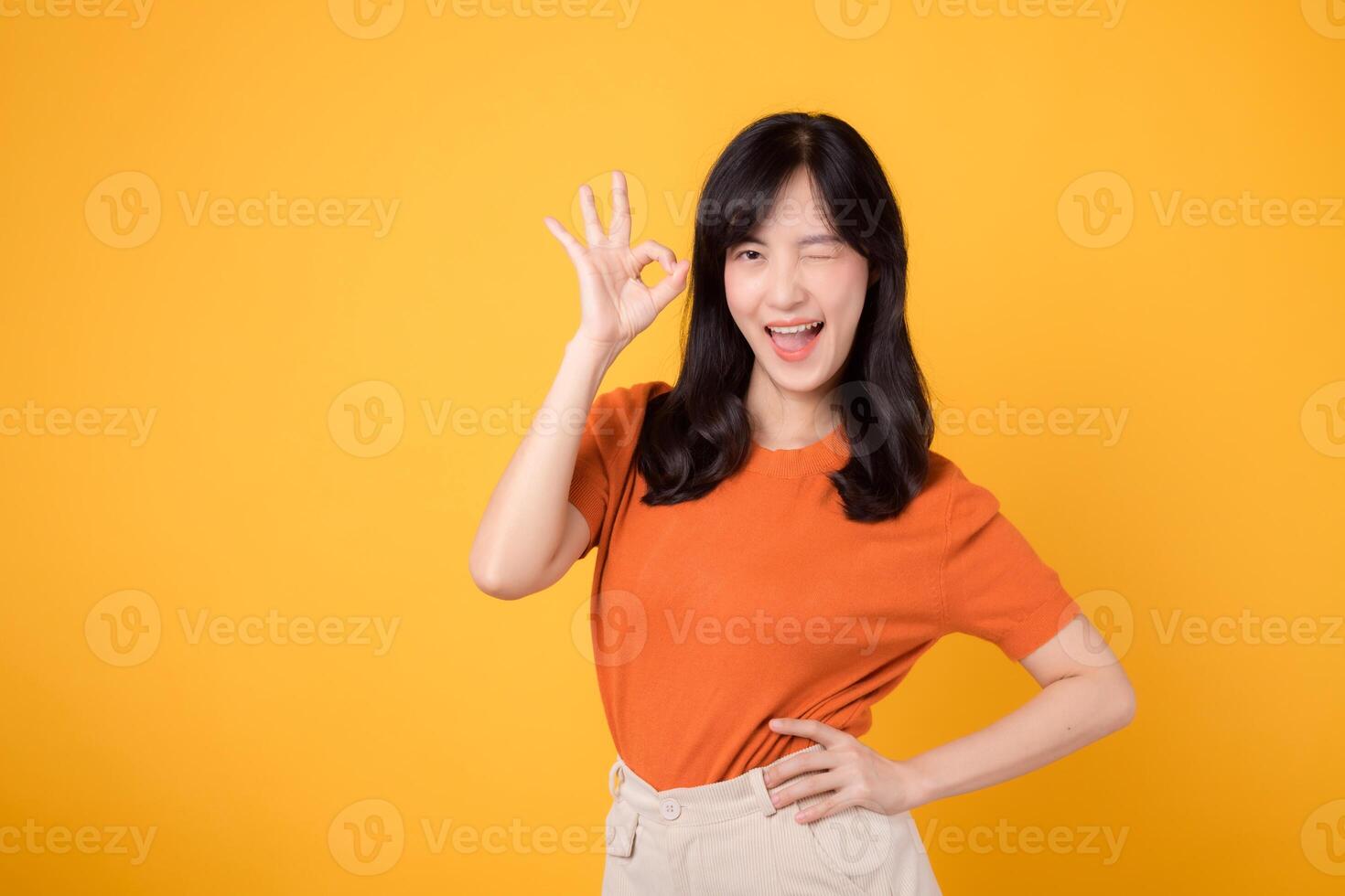 Young asian woman 30s wearing orange shirt showing okay sign gesture isolated on yellow background. Female person with hands gesture concept. photo