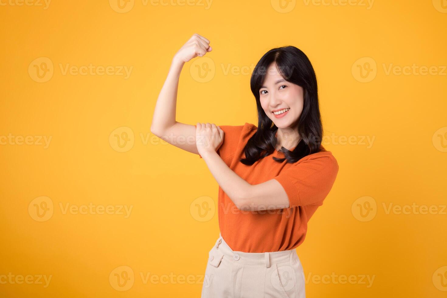 Celebrate with confidence as a young Asian woman in her 30s showcases a fist up hand sign gesture, wearing an orange shirt on yellow background. Empowerment and feminism concept. photo