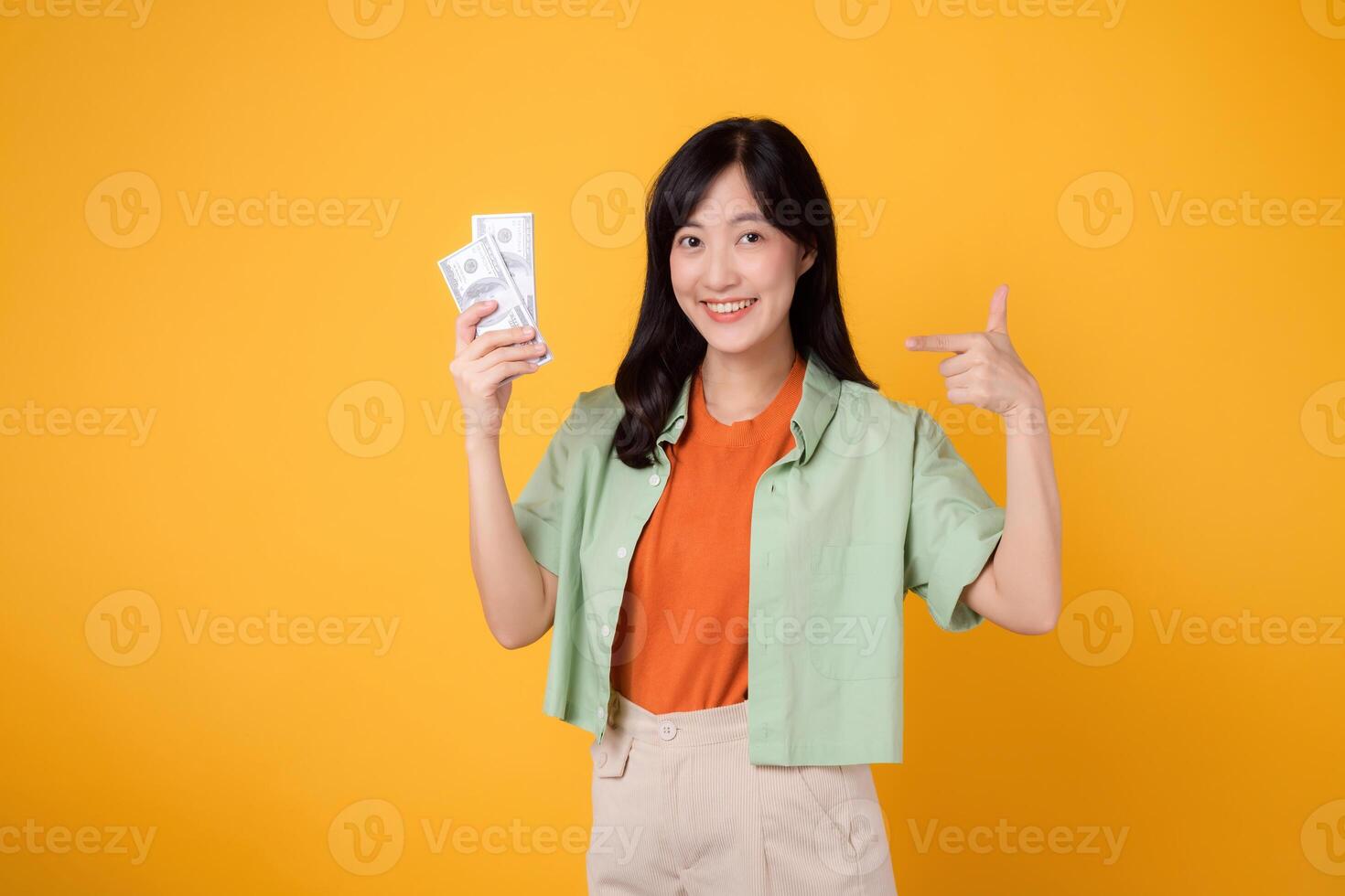 young 30s asian woman happy face dressed in orange shirt and green jumper pointing finger to dollar currency isolated on yellow background. finance business concept. photo