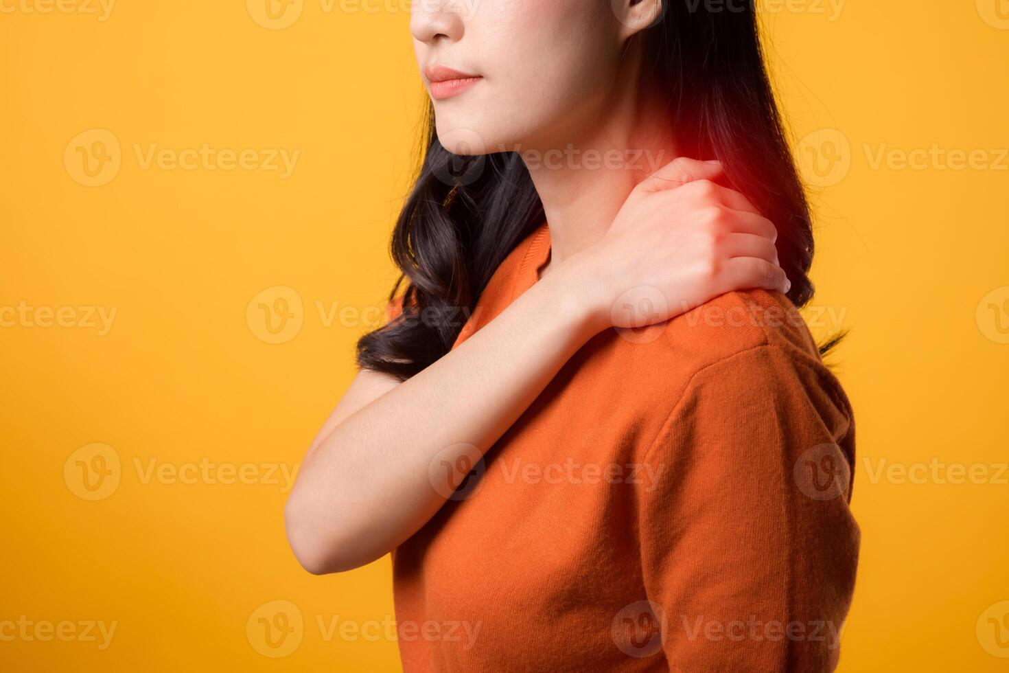 Empathetic young Asian woman in her 30s, wearing an orange shirt, holds her pain shoulder on yellow background. Neck ache therapy medical office syndrome concept. photo