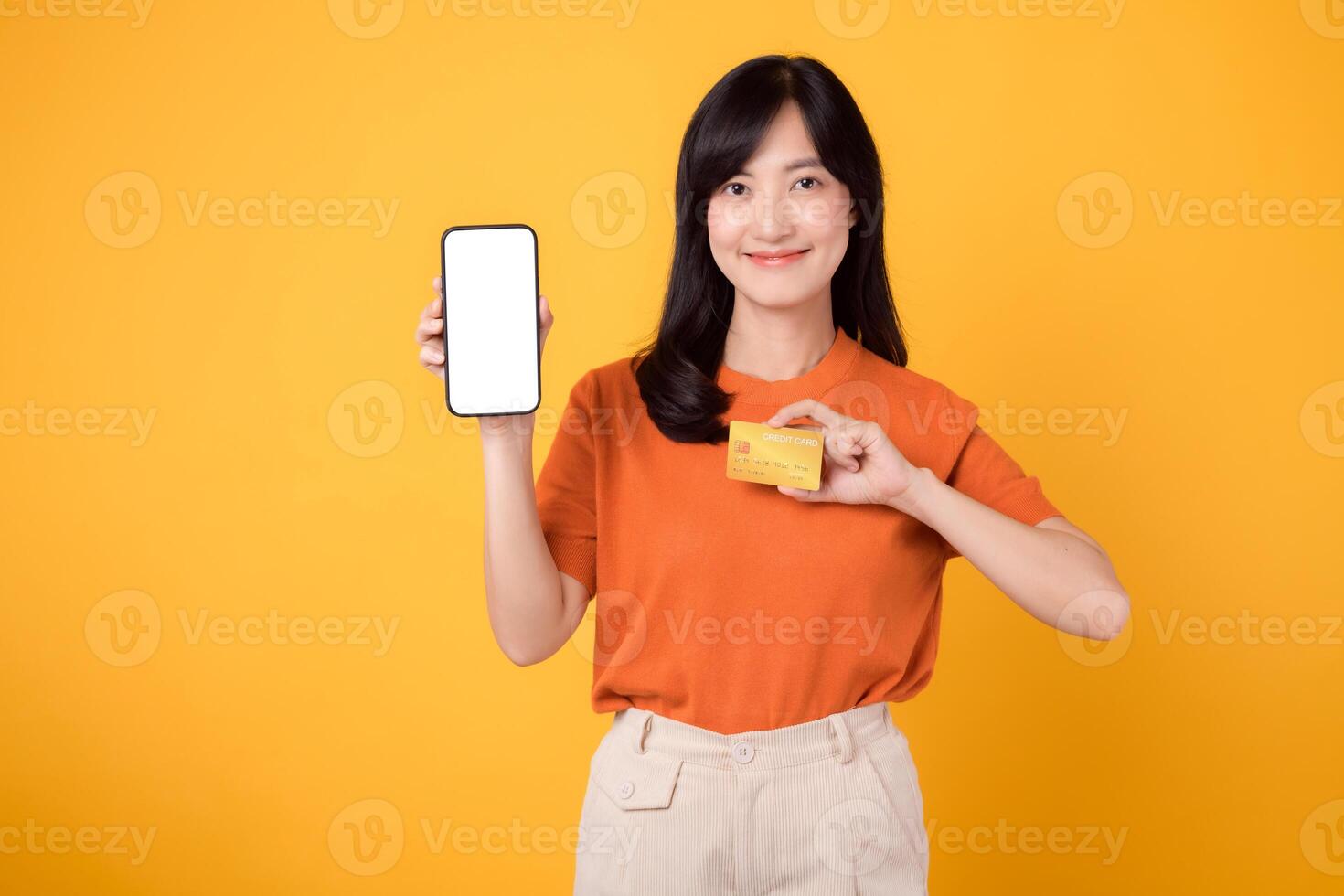 Smiling Asian woman in her 30s, wearing orange shirt, using smartphone, pointing to free copy space on vibrant yellow background. New mobile app concept. photo
