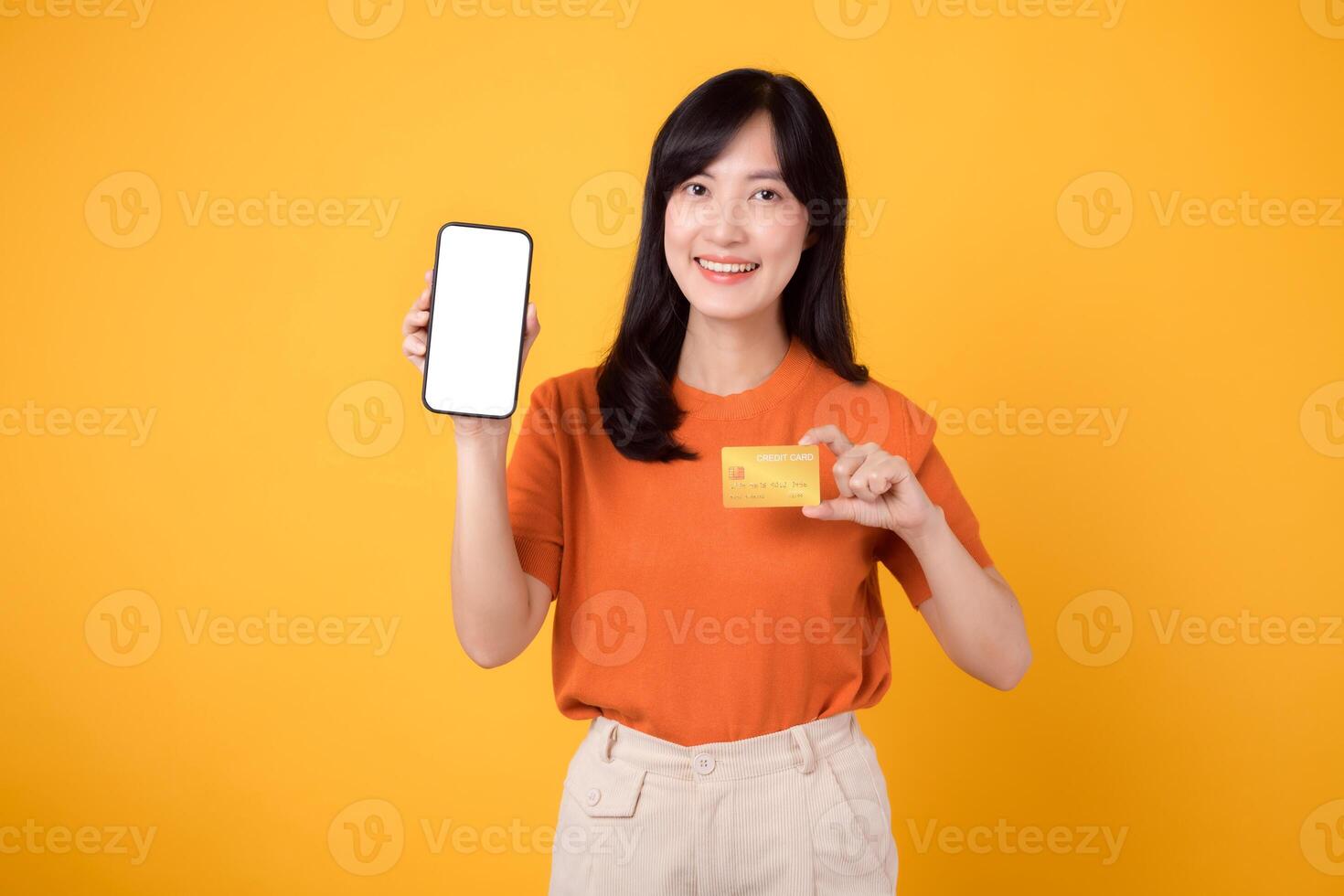 Cheerful Asian woman in her 30s, using smartphone and credit card on yellow background. Streamlined online payment shopping. photo