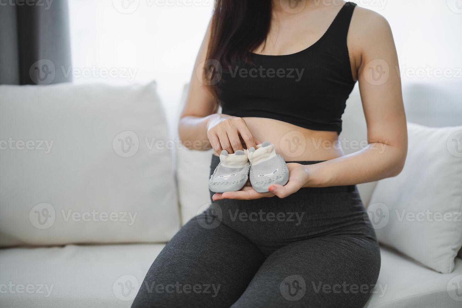 embarazada mujer participación bebé Zapatos en su barriga sentado en sofá sofá. familia madre mamá embarazada concepto. foto