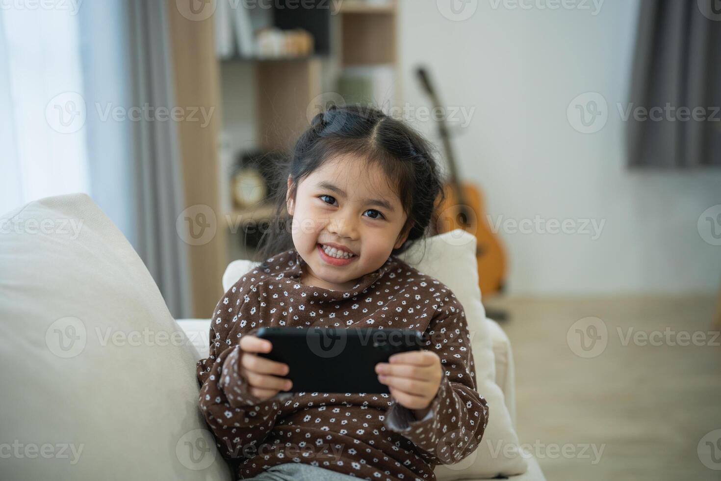 Asian child girl looking using and touch mobile phone screen on couch sofa. Baby smiling funny time to use mobile phone. Too much screen time. Cute girl watching videos while tv, Internet addiction. photo